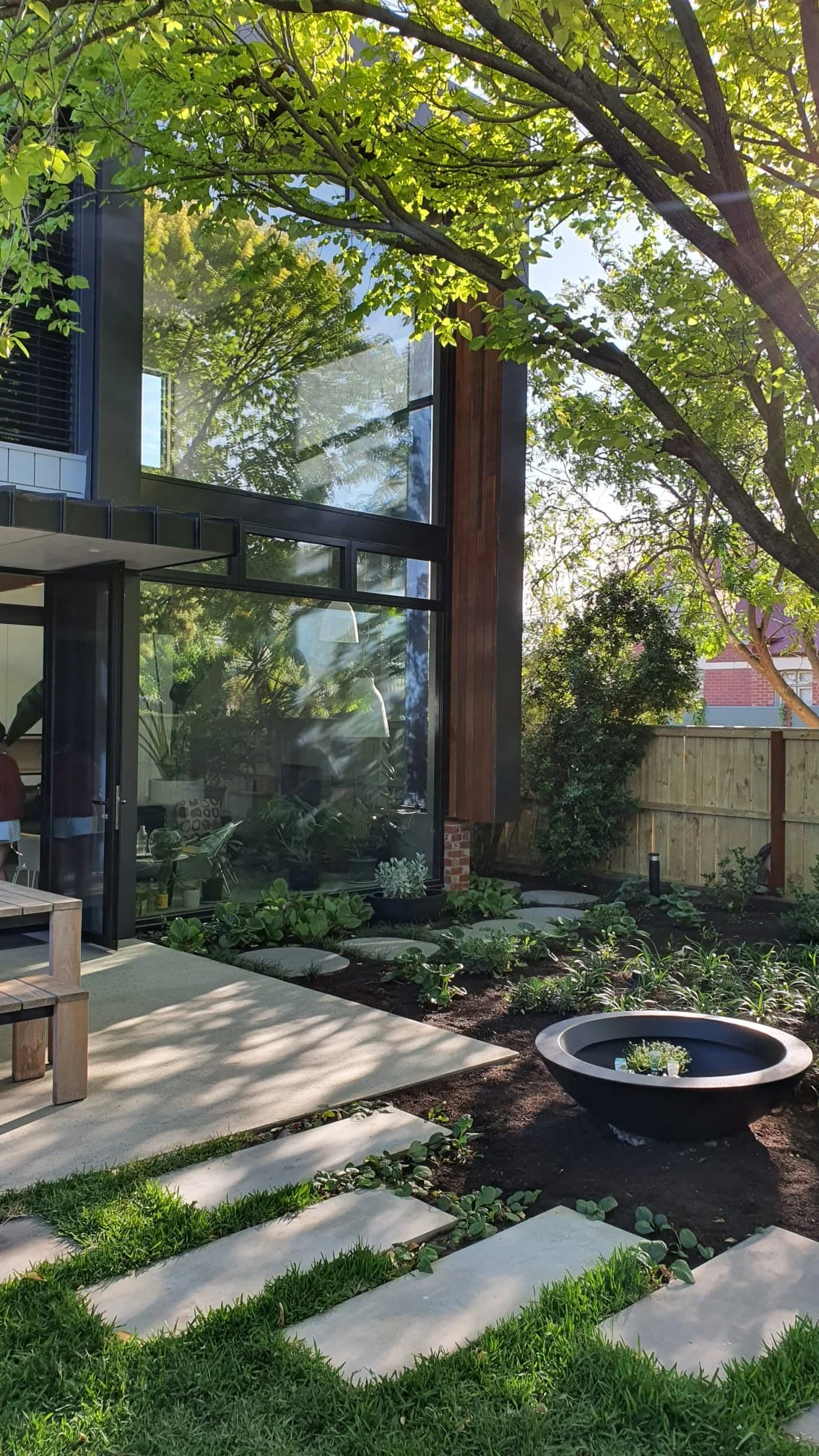 A modern house with large glass windows, surrounded by lush green trees and plants in a garden. There is a patio with a small wooden table and shelves, and a garden bed with leafy green plants. A black round water feature is also visible.