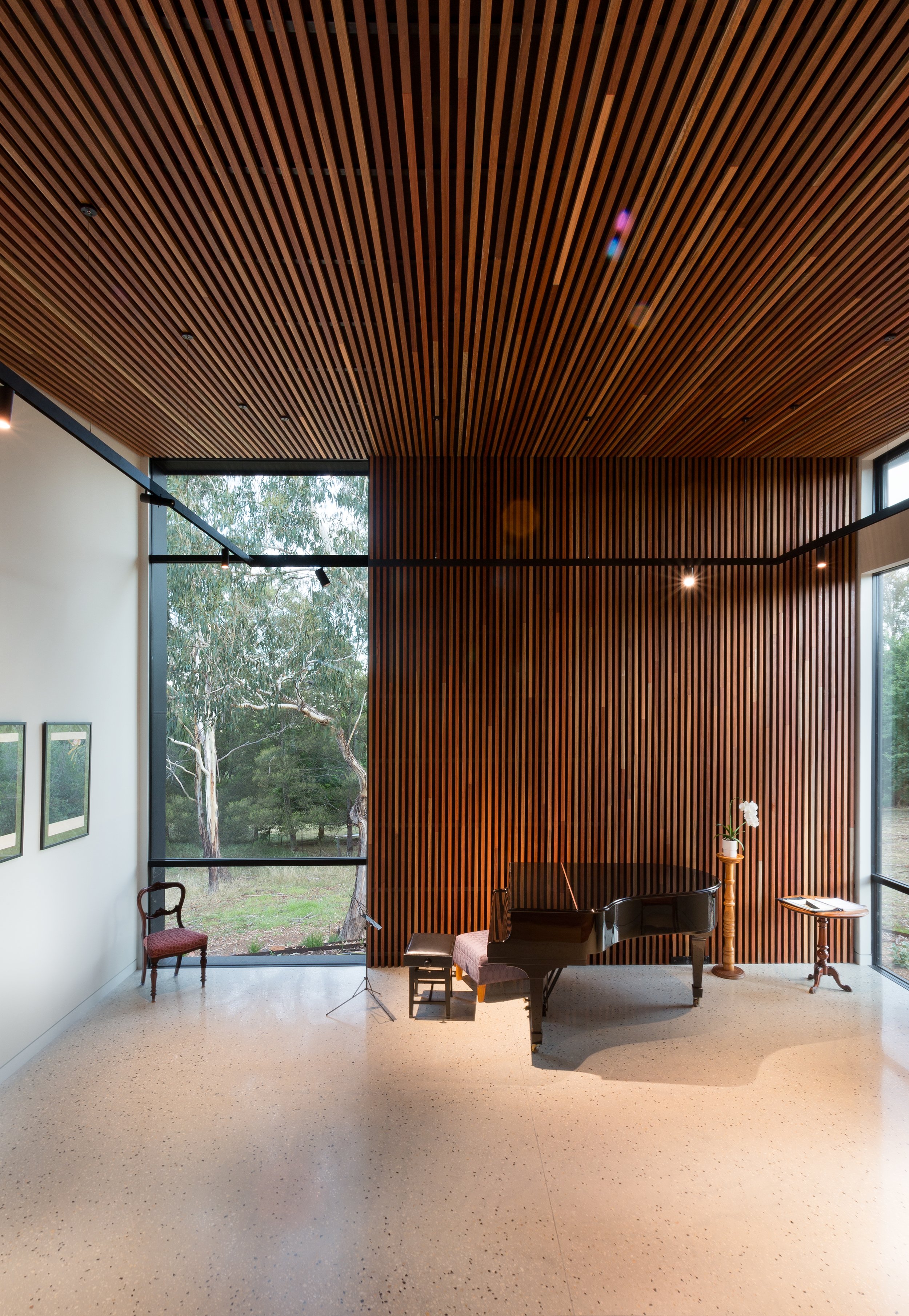 Interior view of a room with a high wooden slat ceiling and wall, large windows showing trees outside, a black grand piano, and a small table with a potted white orchid.