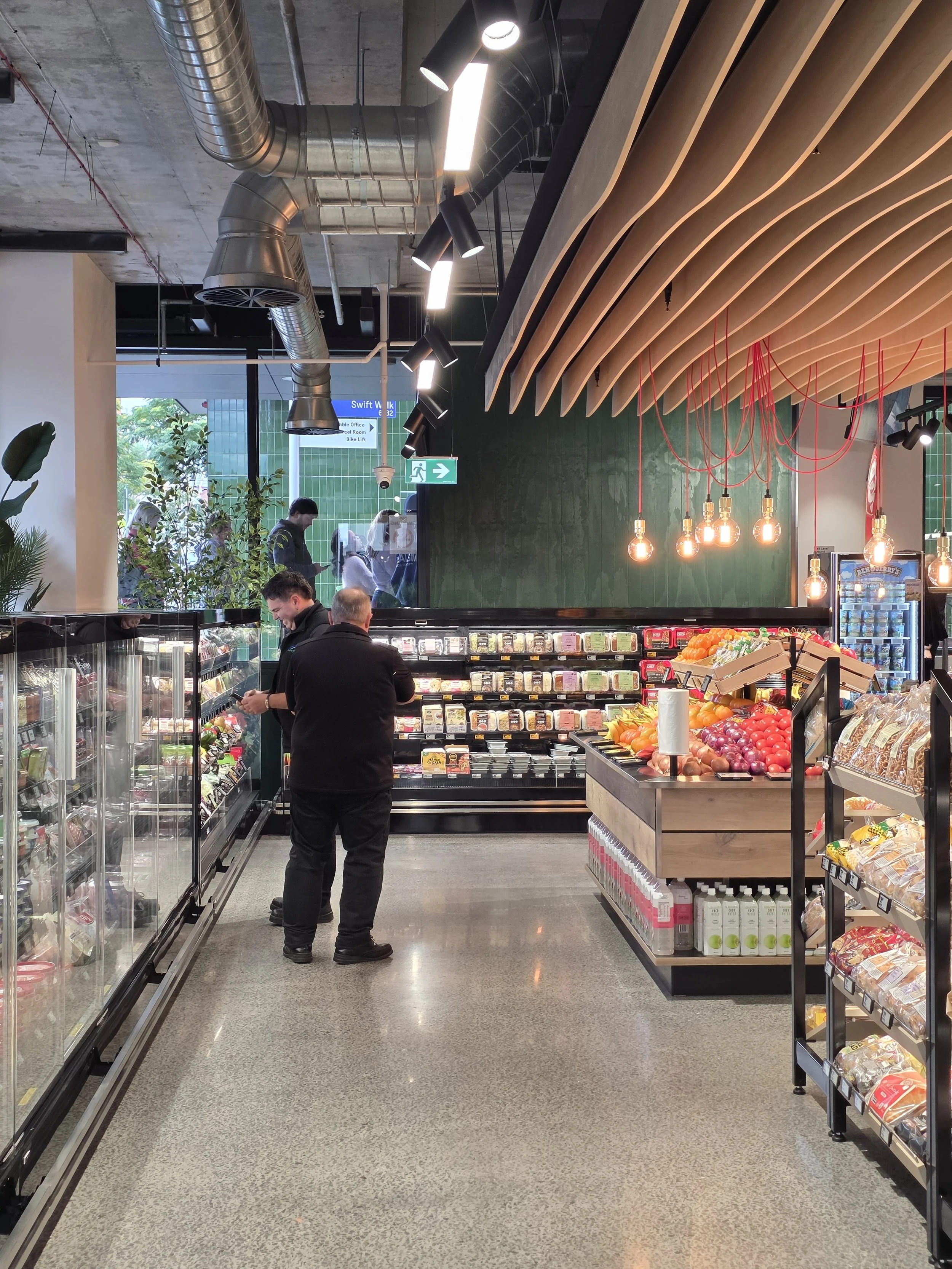 Inside a grocery store, two men are shopping near refrigerated sections stocked with various packaged foods and fresh produce, with modern lighting and ceiling ductwork visible.