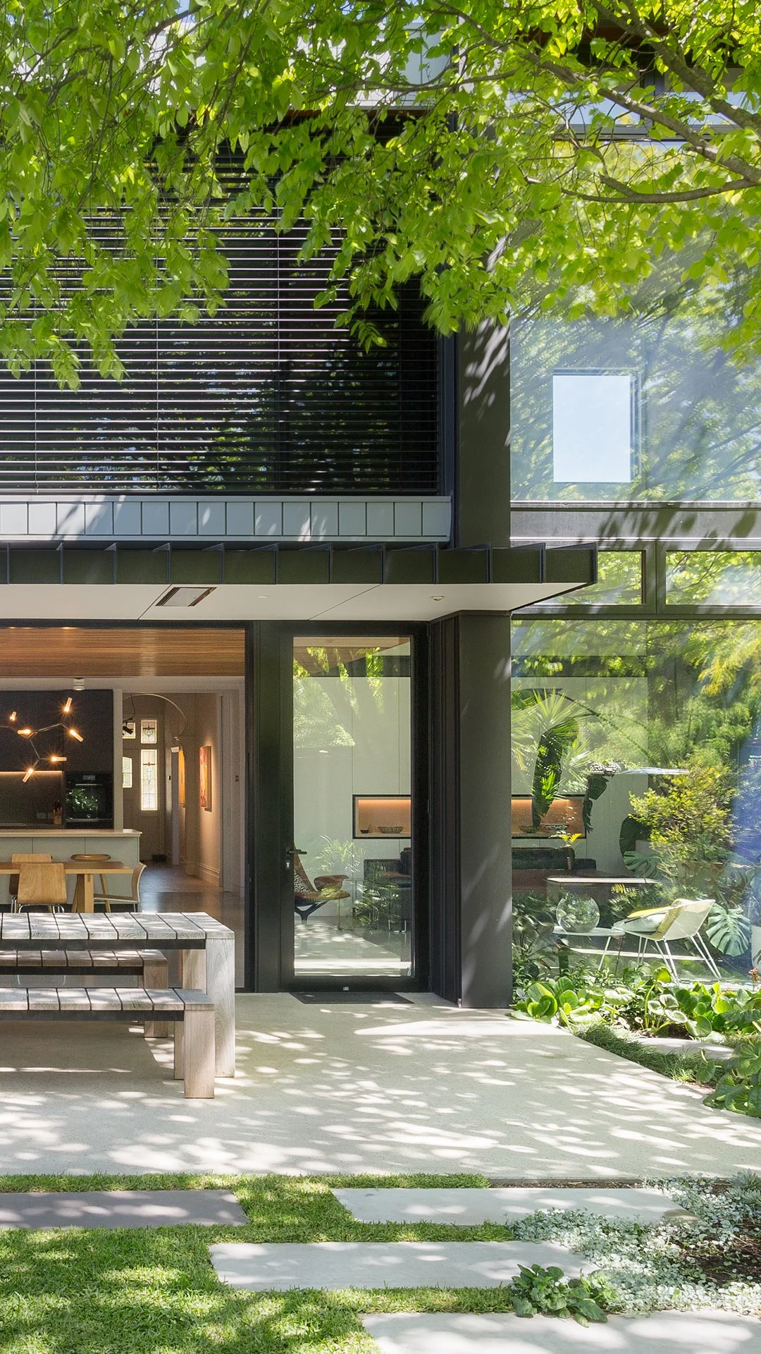 Modern house with large glass windows, surrounded by green trees and lush garden, with outdoor seating and pathway.