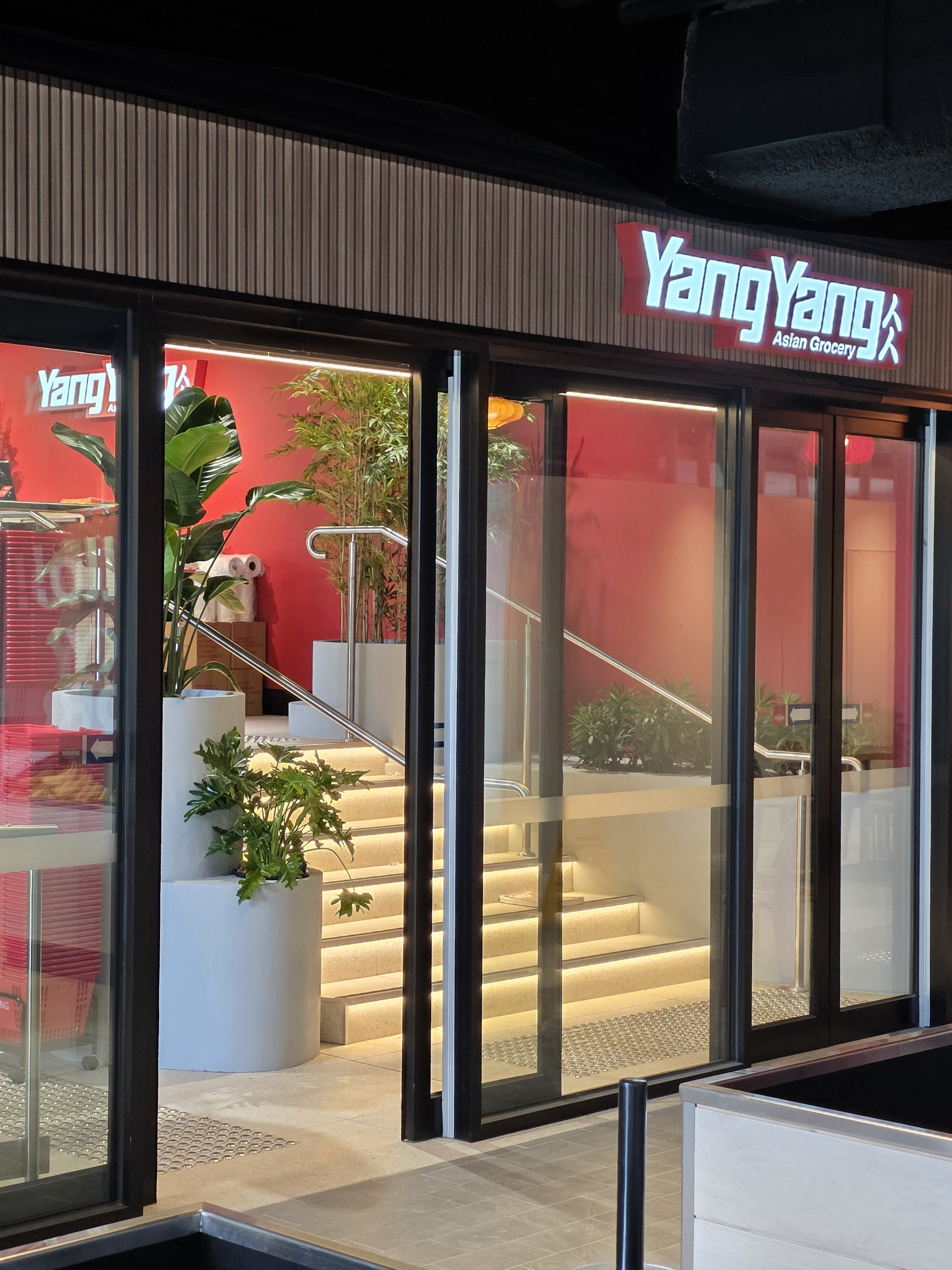 The entrance of Yang Yang Asian Grocery, with glass doors, indoor plants, stairs, and illuminated signage.