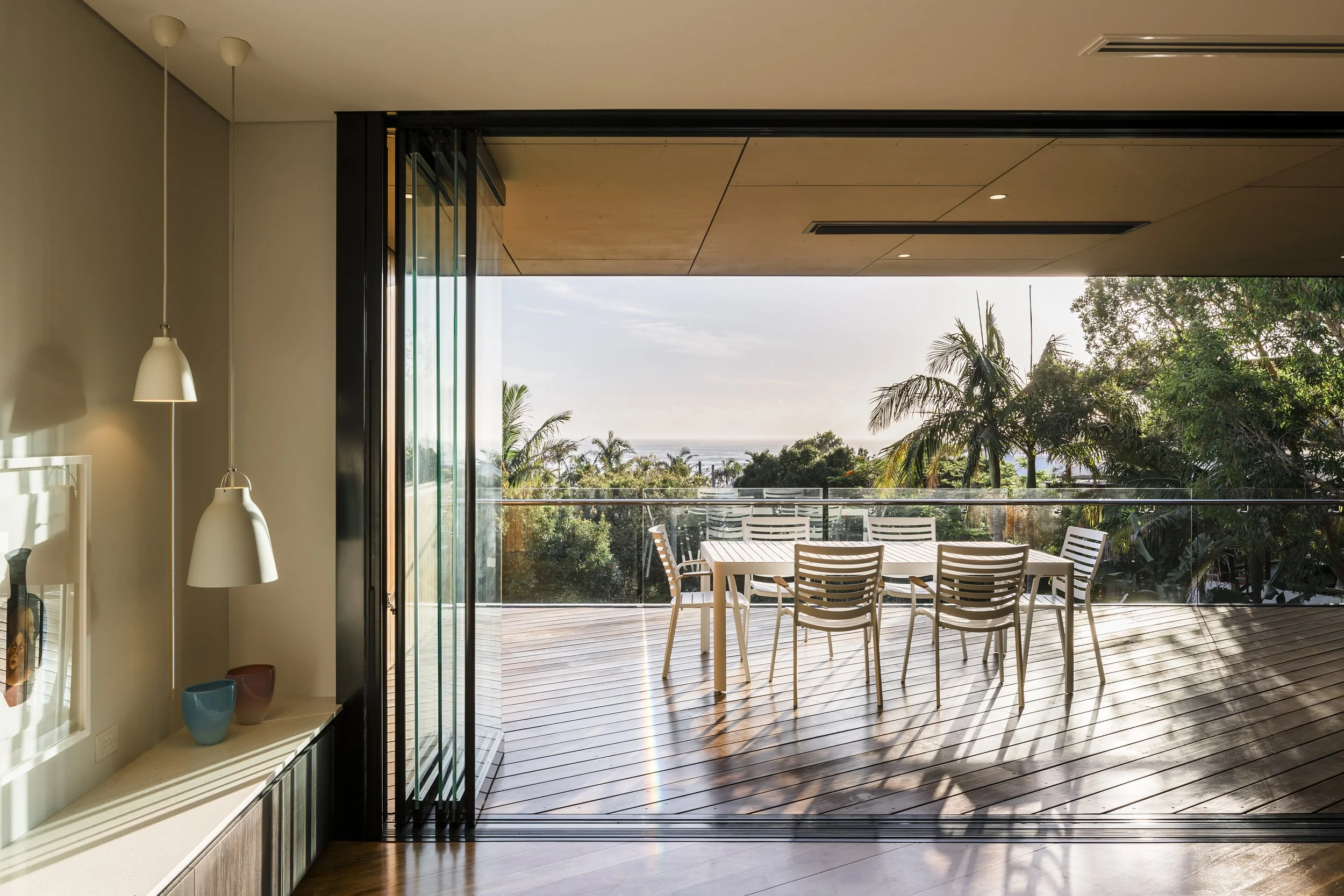 Open balcony door leading to an outdoor dining area with a long rectangular table and multiple chairs, overlooking trees and the ocean in the distance. Indoor area has a ceiling with embedded lighting and a shelf with decorative vases, with sunlight 