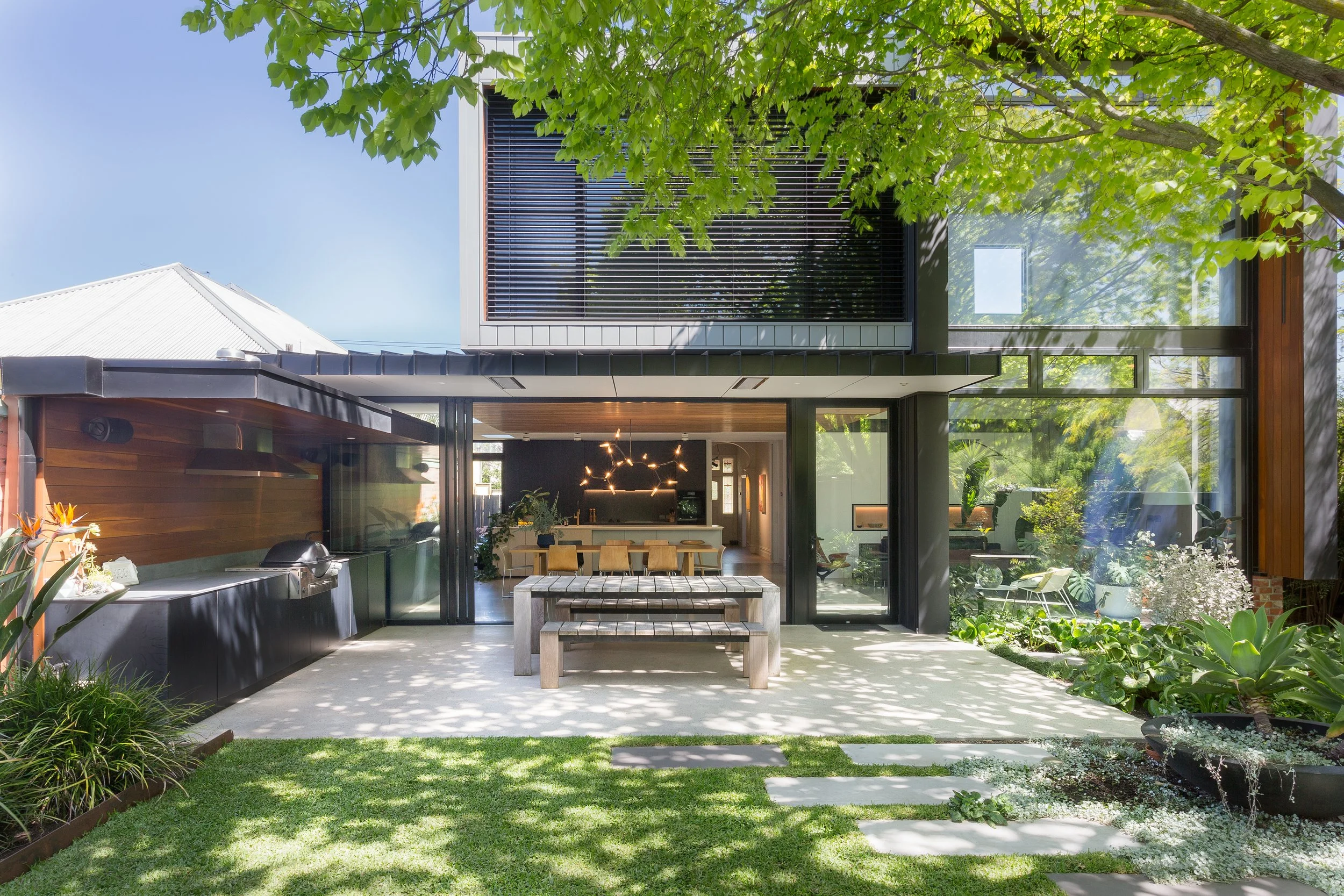 Modern house with large glass windows, a backyard garden with green grass, plants, and a patio area with a wooden dining table and benches, under a tree with lush foliage.
