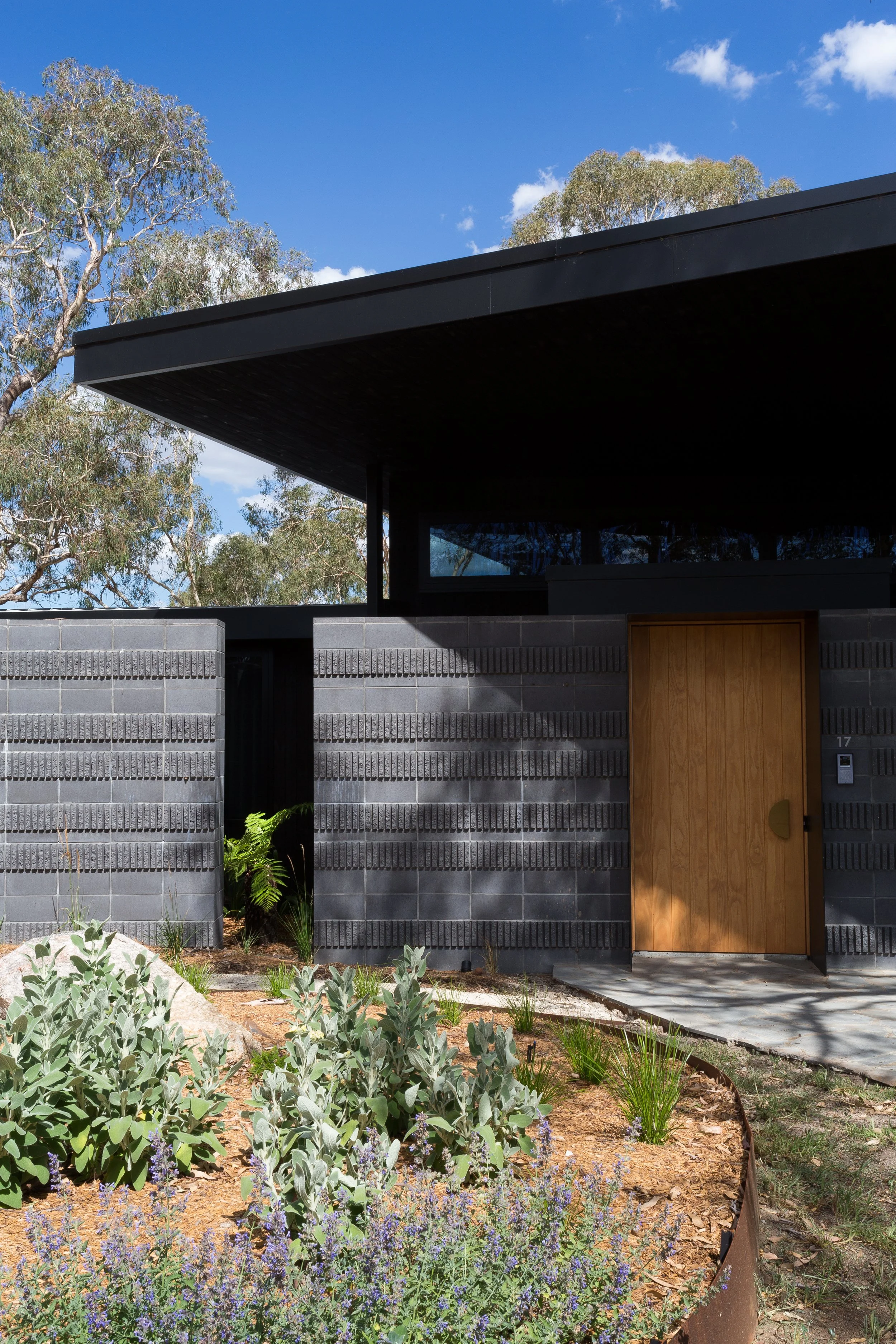 Modern house with black roof, black brick wall, and wooden door, surrounded by a garden with succulents and flowering plants under a blue sky with scattered clouds.