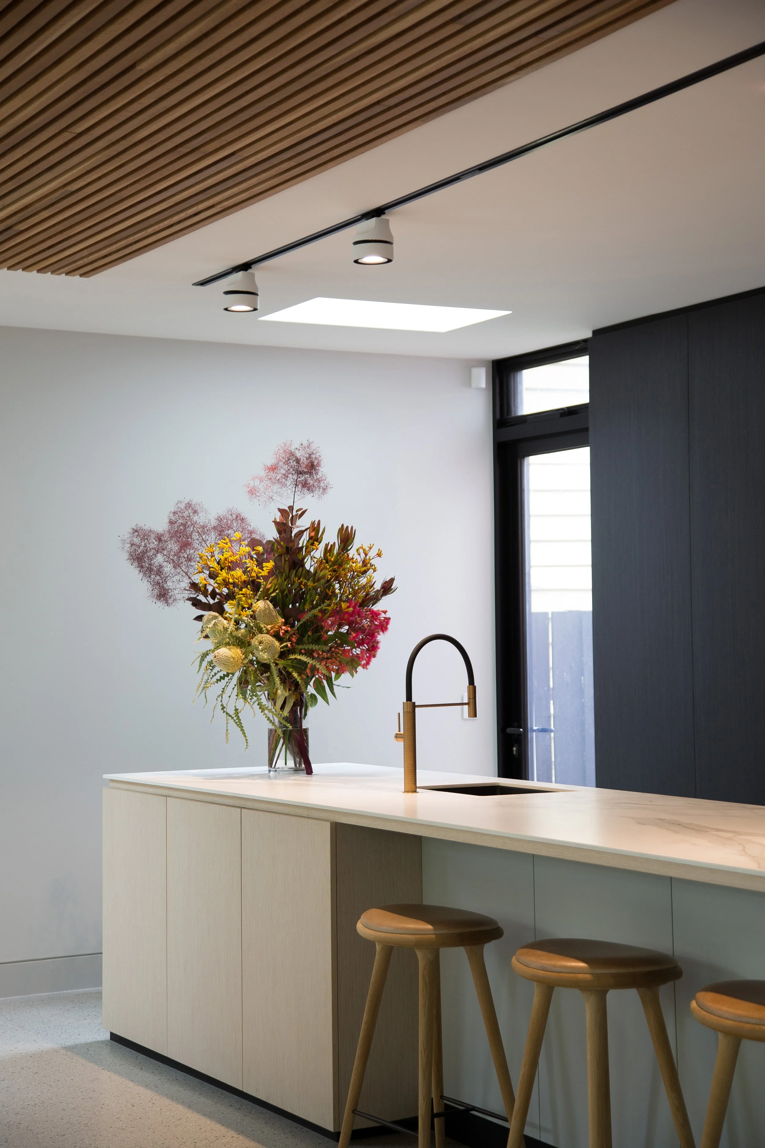 Modern kitchen with a white island, a tall floral arrangement, three wooden barstools, a black wall, a window, and ceiling track lighting.