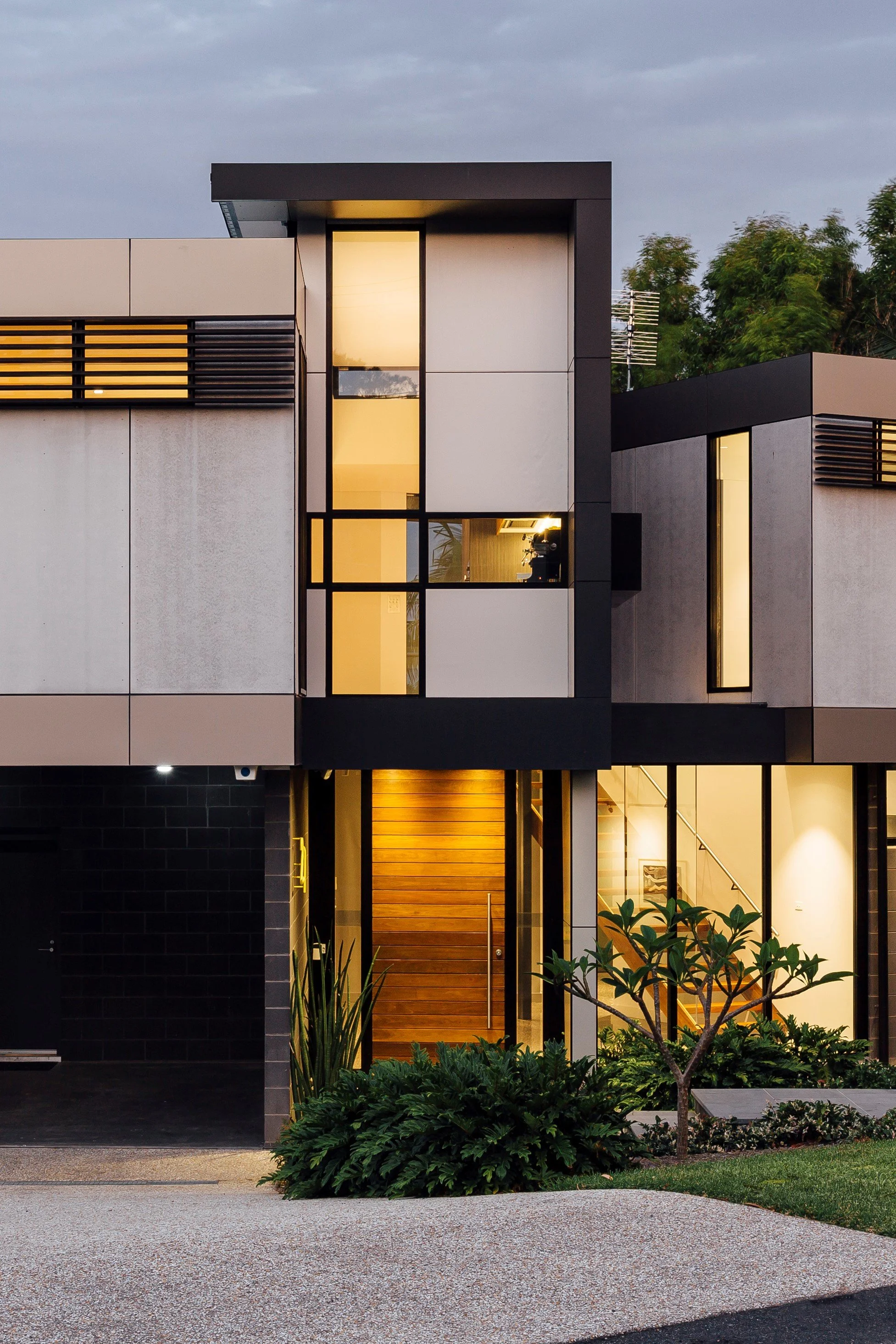 Modern multi-story house with large windows, illuminated interior, wooden entrance door, surrounded by greenery and trees, evening lighting.