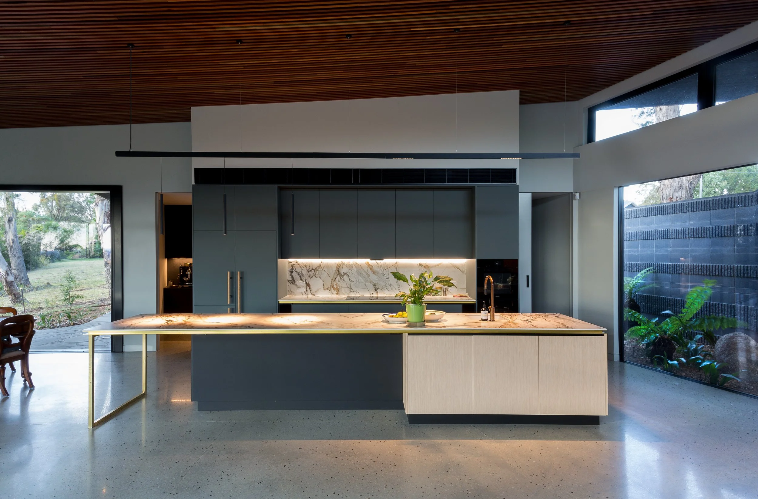 Modern kitchen with an island, marble countertop, black cabinets, and large windows showing an outdoor garden.