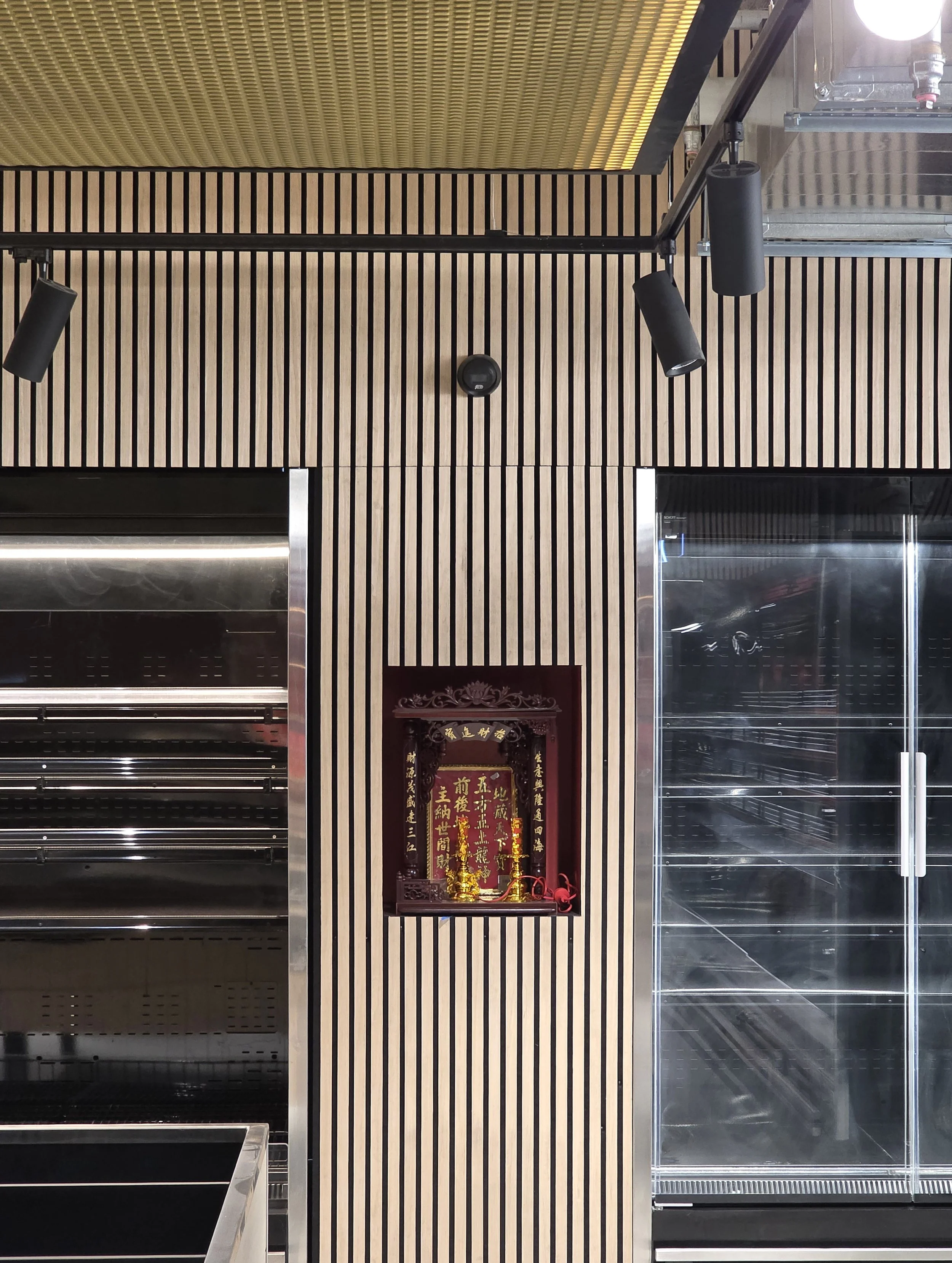 Empty shopping display cases with black and glass shelves, a decorative altar with gold objects and Chinese characters in the center, and a wooden slat wall behind, in a modern retail space.