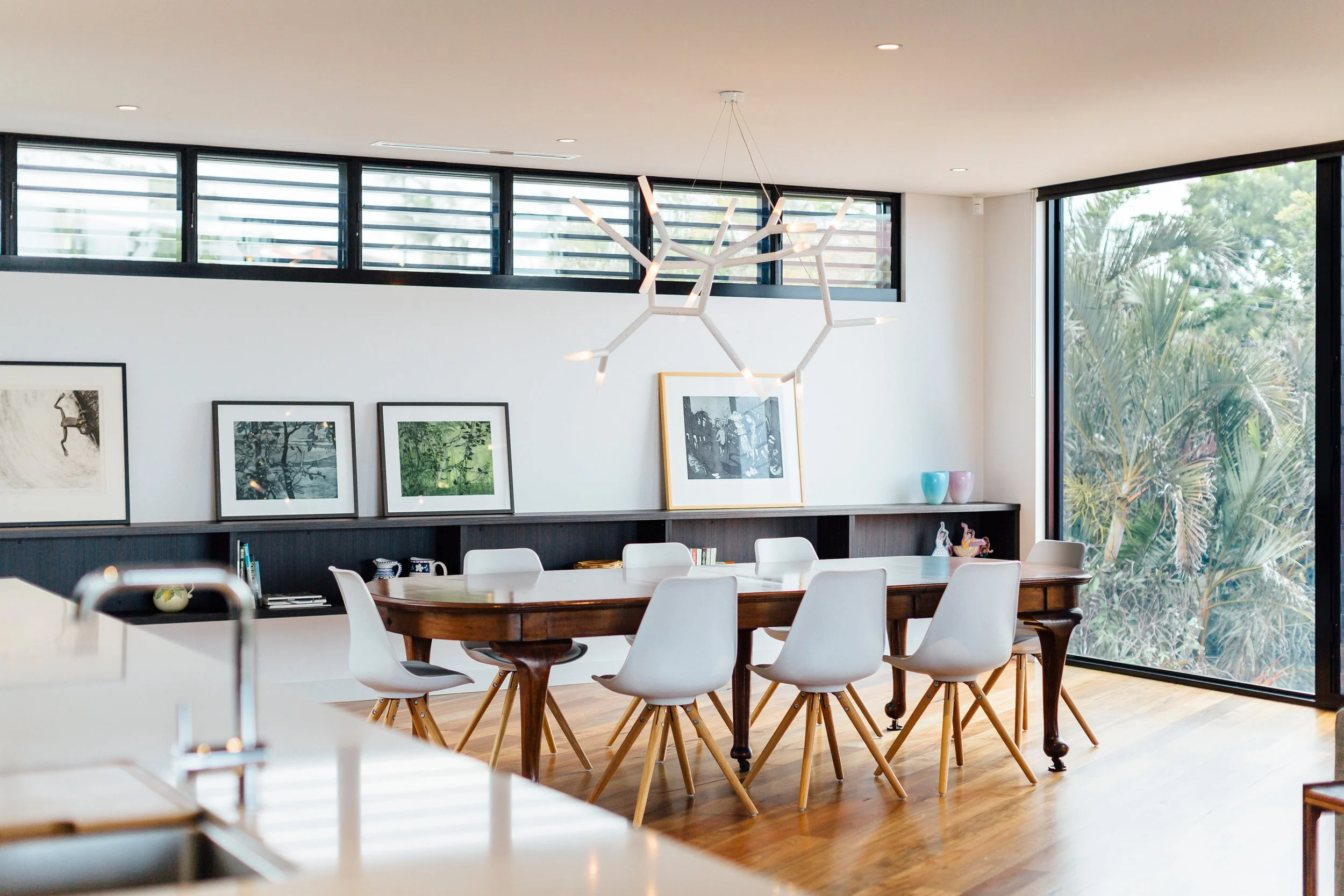 Modern dining room with a large wooden table surrounded by white chairs, minimalist paintings on the wall, and large windows with horizontal blinds and a sliding glass door, allowing natural light and views of greenery outside.