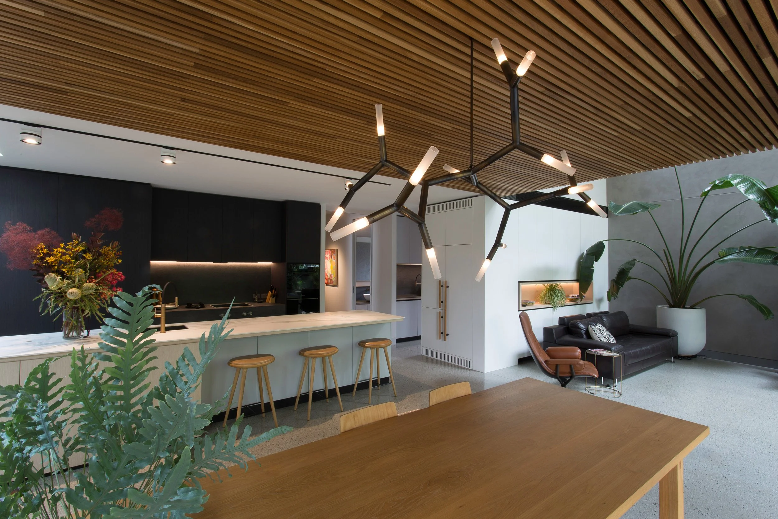 Modern kitchen and living area with wooden ceiling, black and white cabinets, a hanging chandelier, a couch, a chair, and large potted plants.