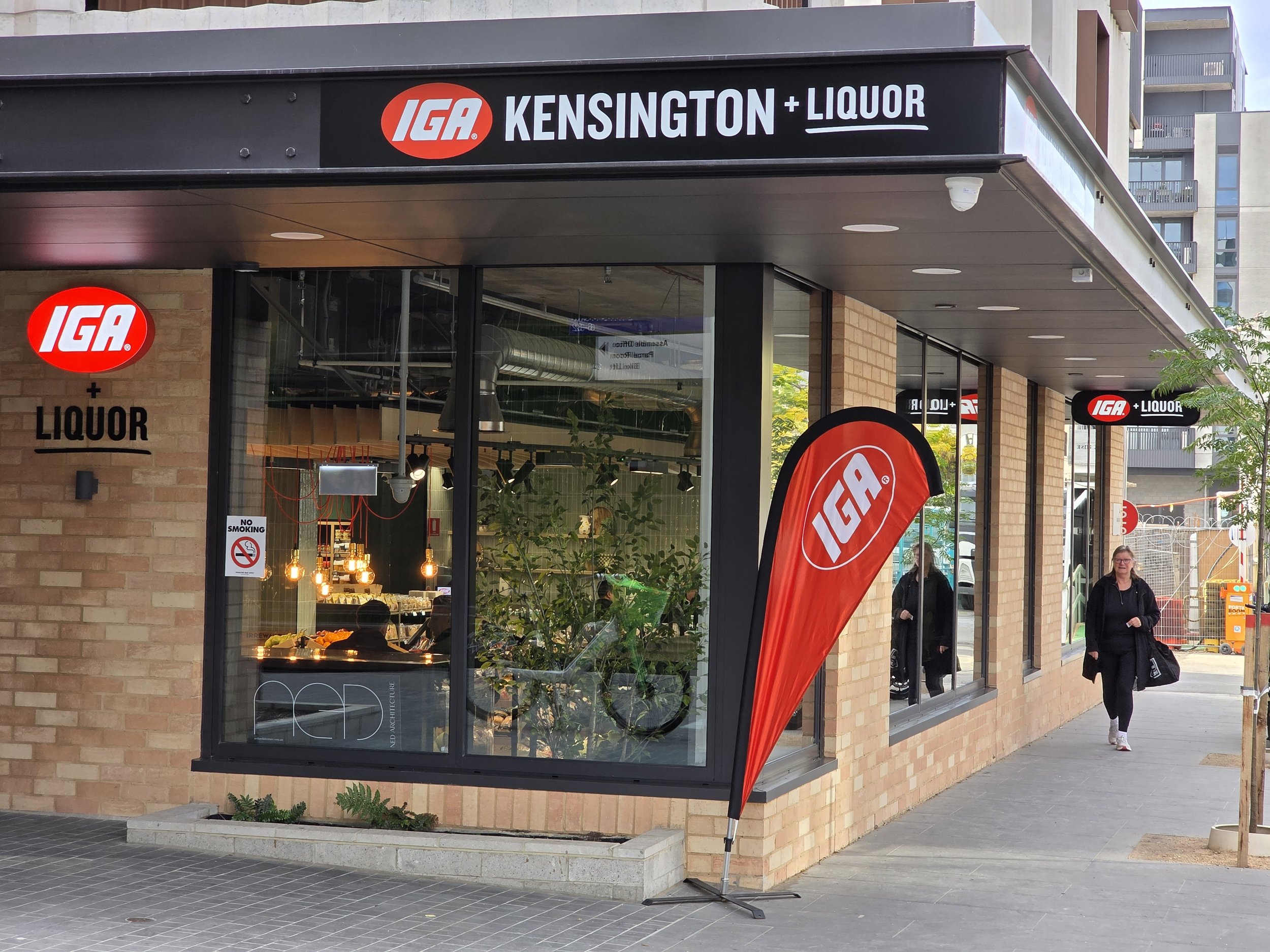 Exterior of an IGA grocery store with signage indicating shopping for Kensington and liquor. The store has large glass windows, and there is a red IGA flag outside. There are people walking on the sidewalk beside the store.