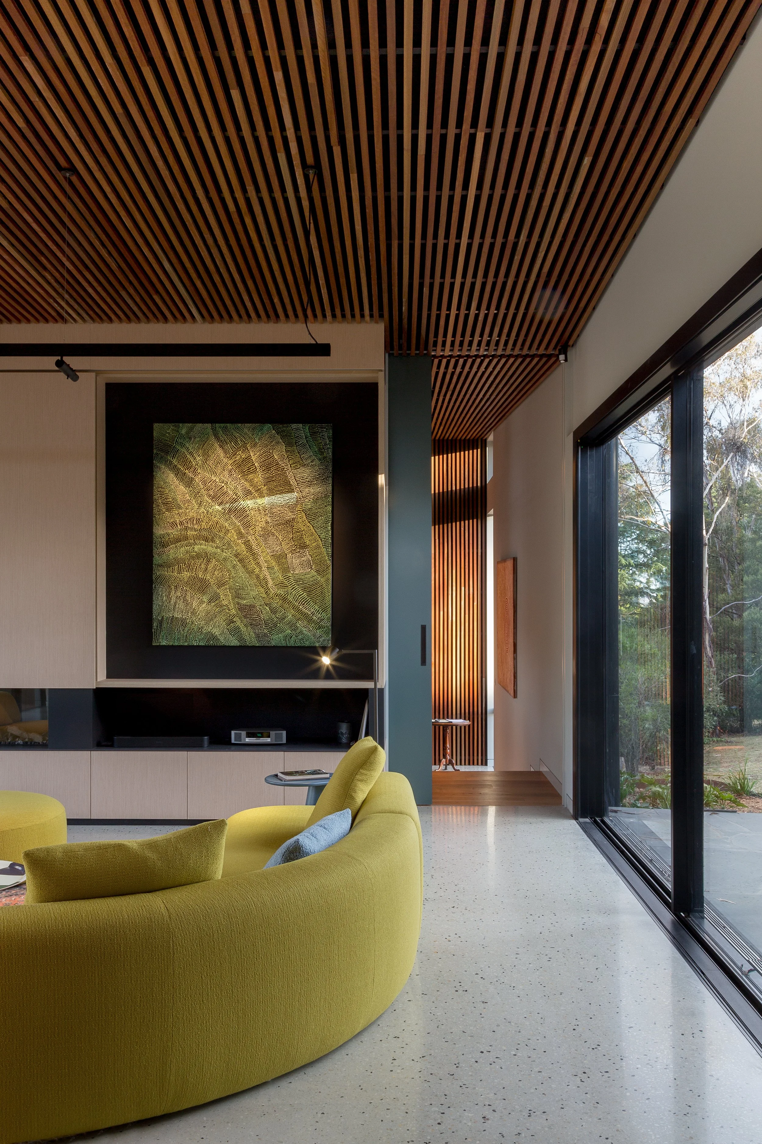Modern living room with a green curved sofa, a flat-screen TV, a large window with black framing, wooden slat ceiling, and an abstract wall art piece.