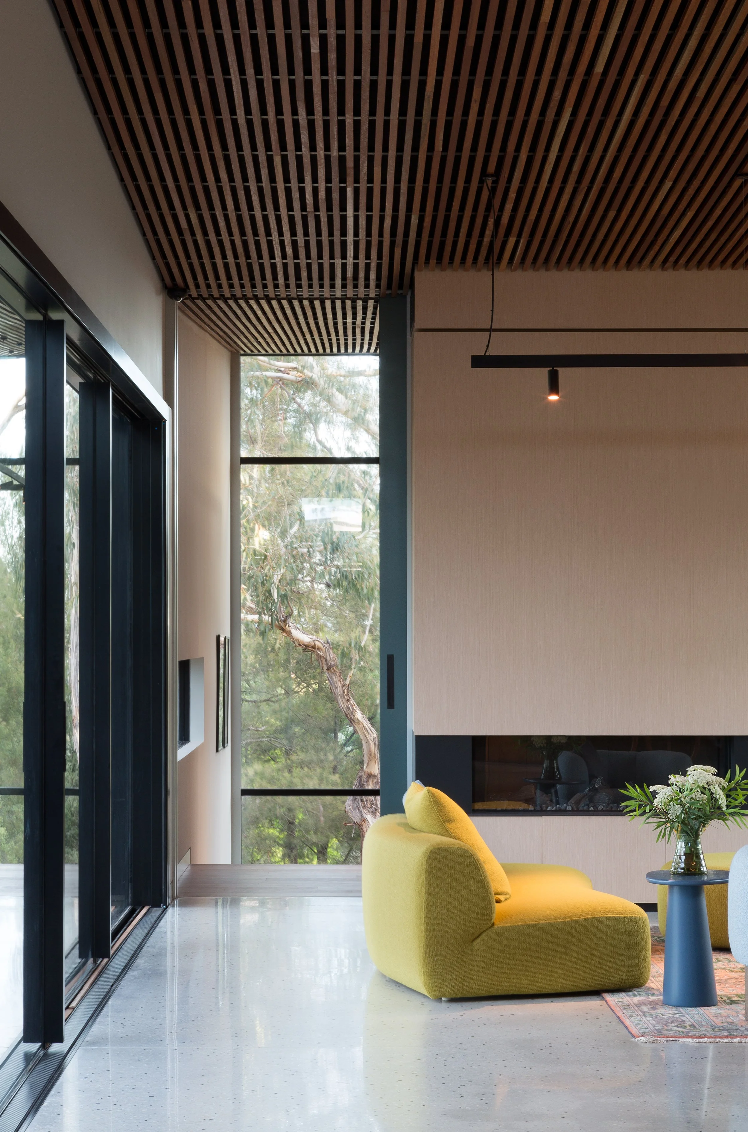 Interior view of a modern living room with large windows, wood slat ceiling, a yellow sofa, blue side table, and a fireplace, with trees visible outside.