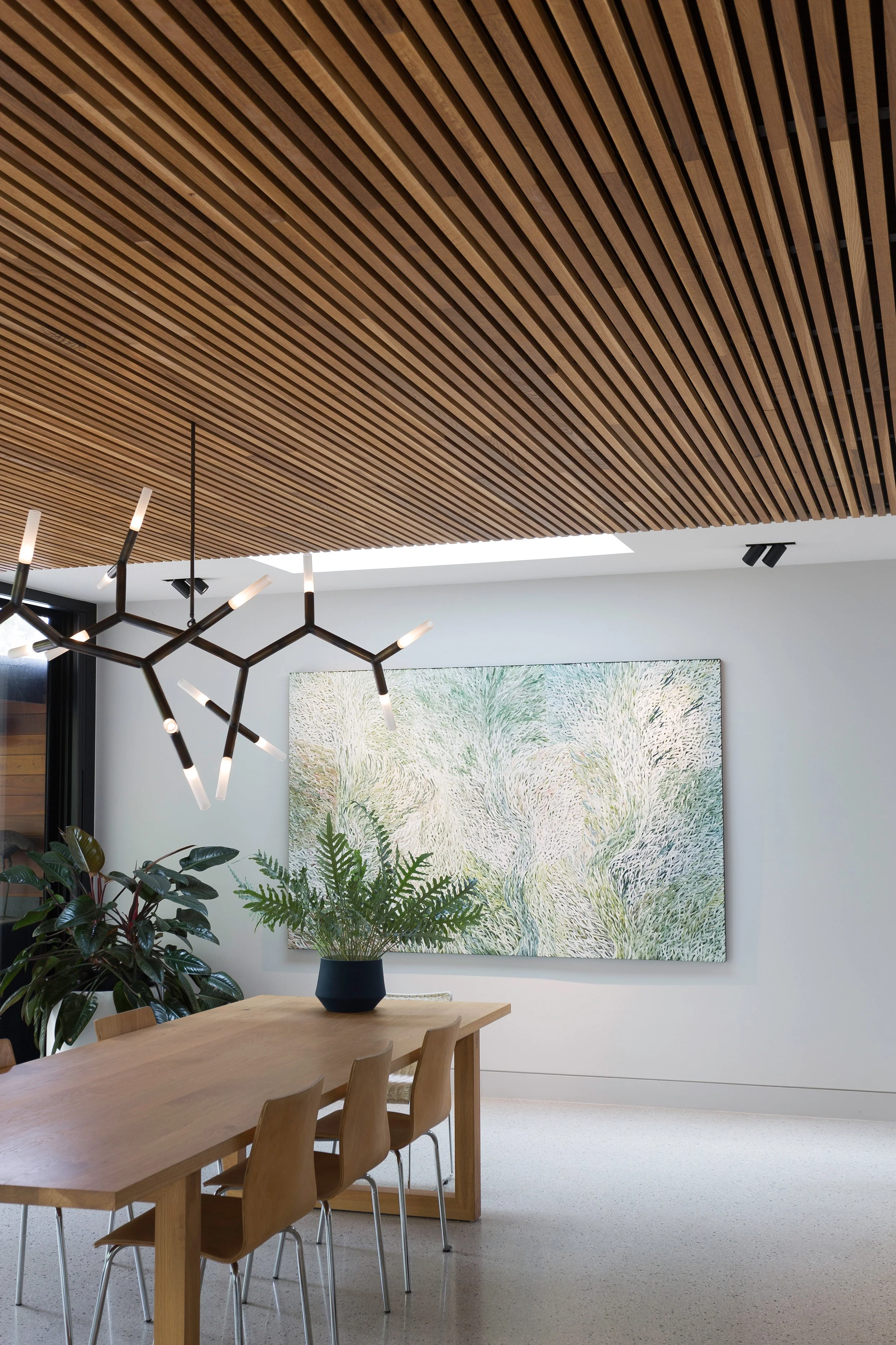 Modern dining room with wooden table, wooden chairs, indoor plants, abstract wall art, and a wooden slat ceiling with hanging light fixture.