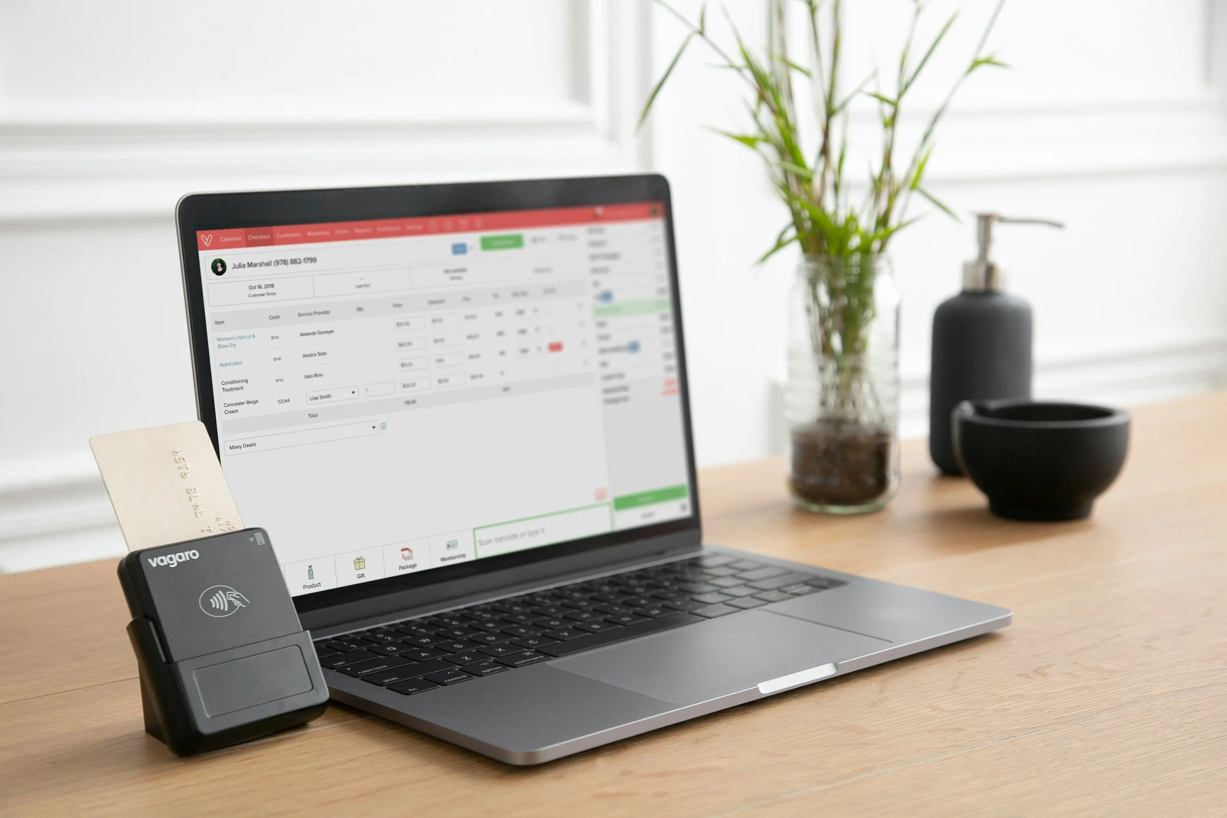 A laptop on a wooden desk displaying a business software interface, with a security scanner device and a credit card in the foreground, and a glass vase with plant and black container in the background.