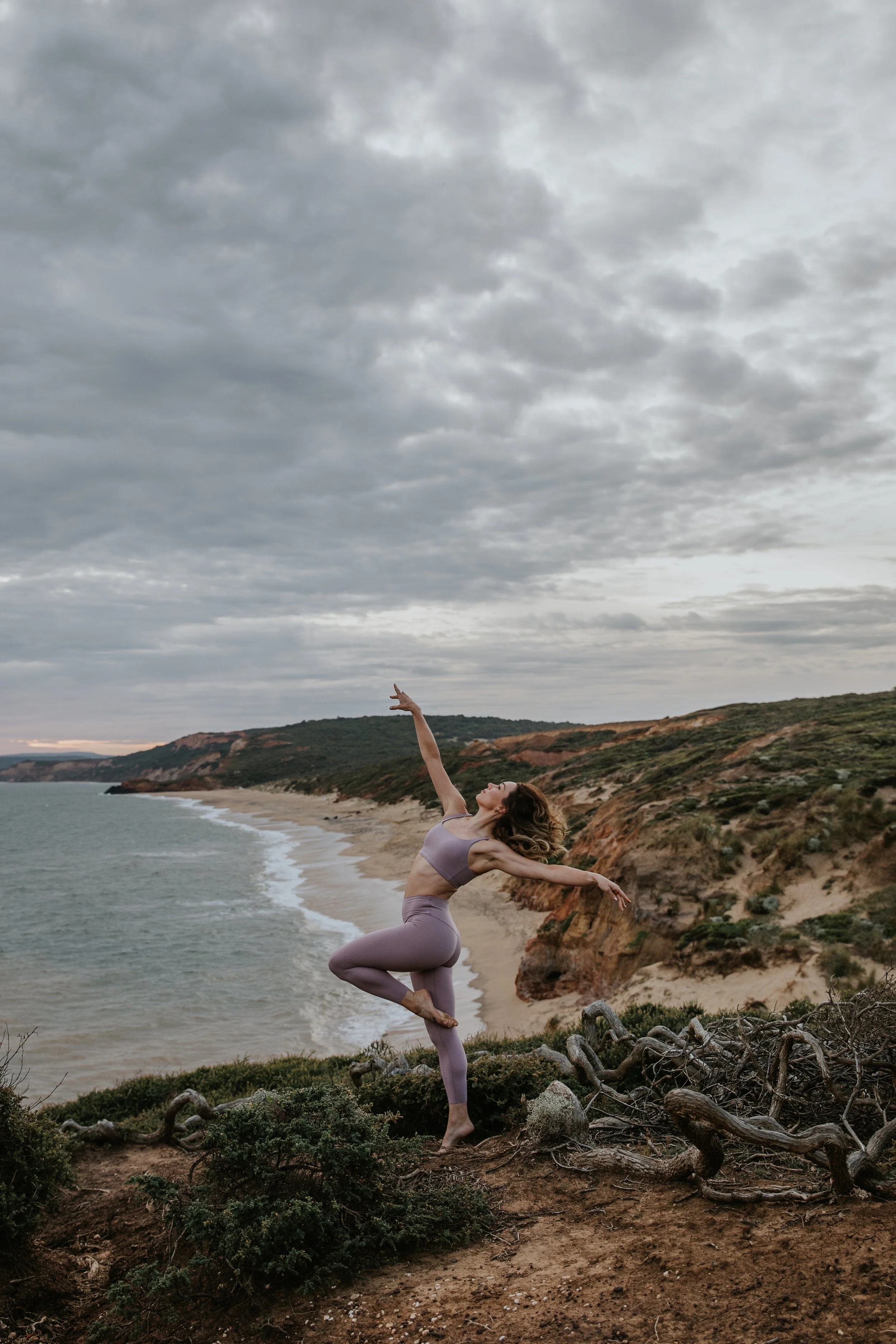 Stef Wild athletic wear posing gracefully on a coastal cliff with ocean and cloudy sky in the background.