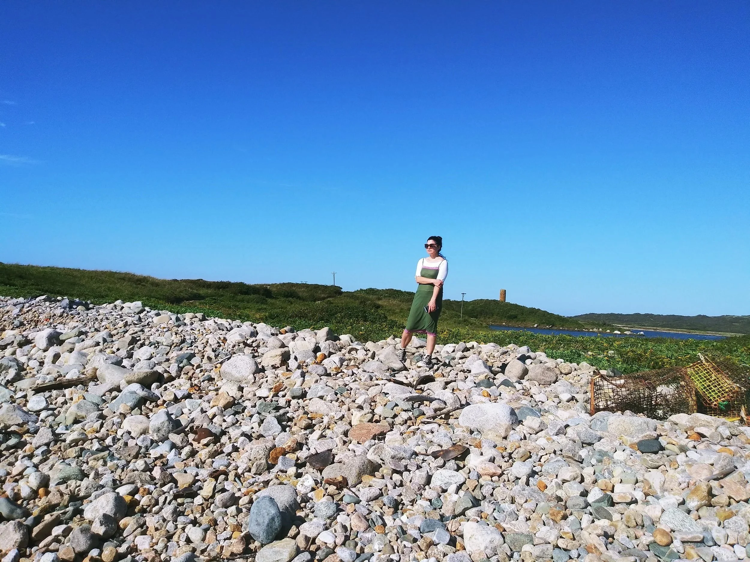 Photo of woman in sunglasses standing on rocky beach.