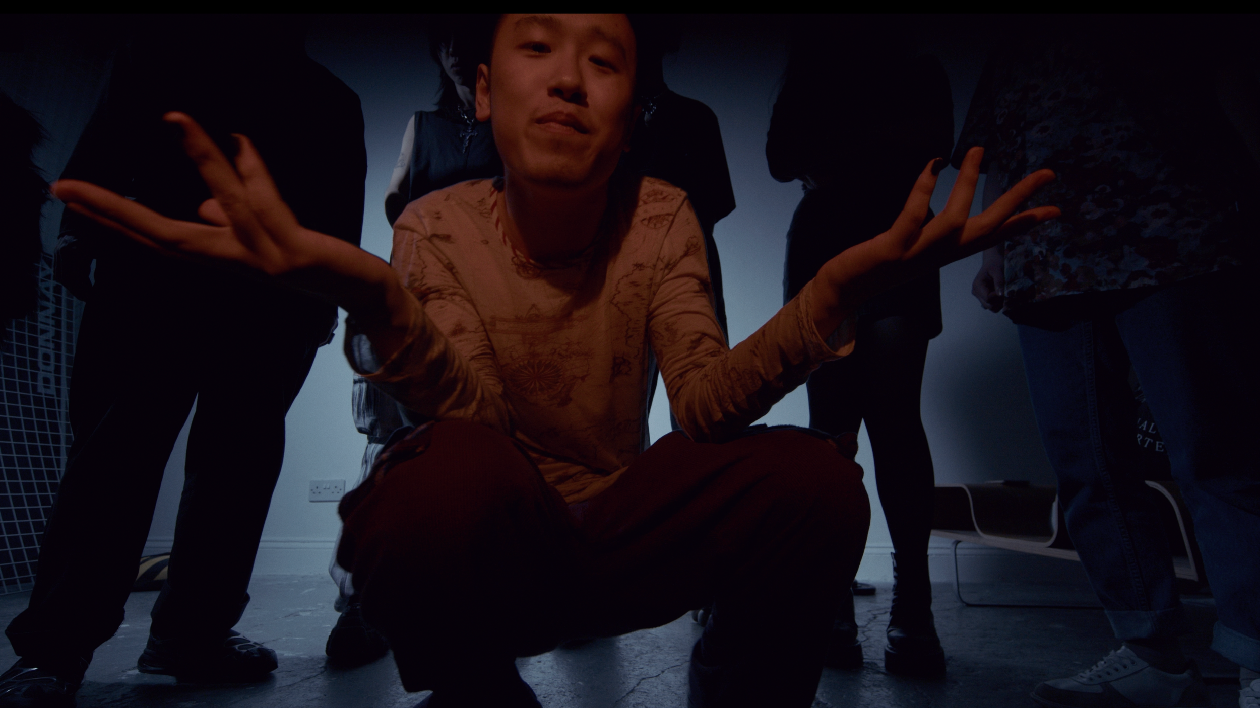 Person crouching in the center with arms extended, surrounded by other people in a dimly lit room.