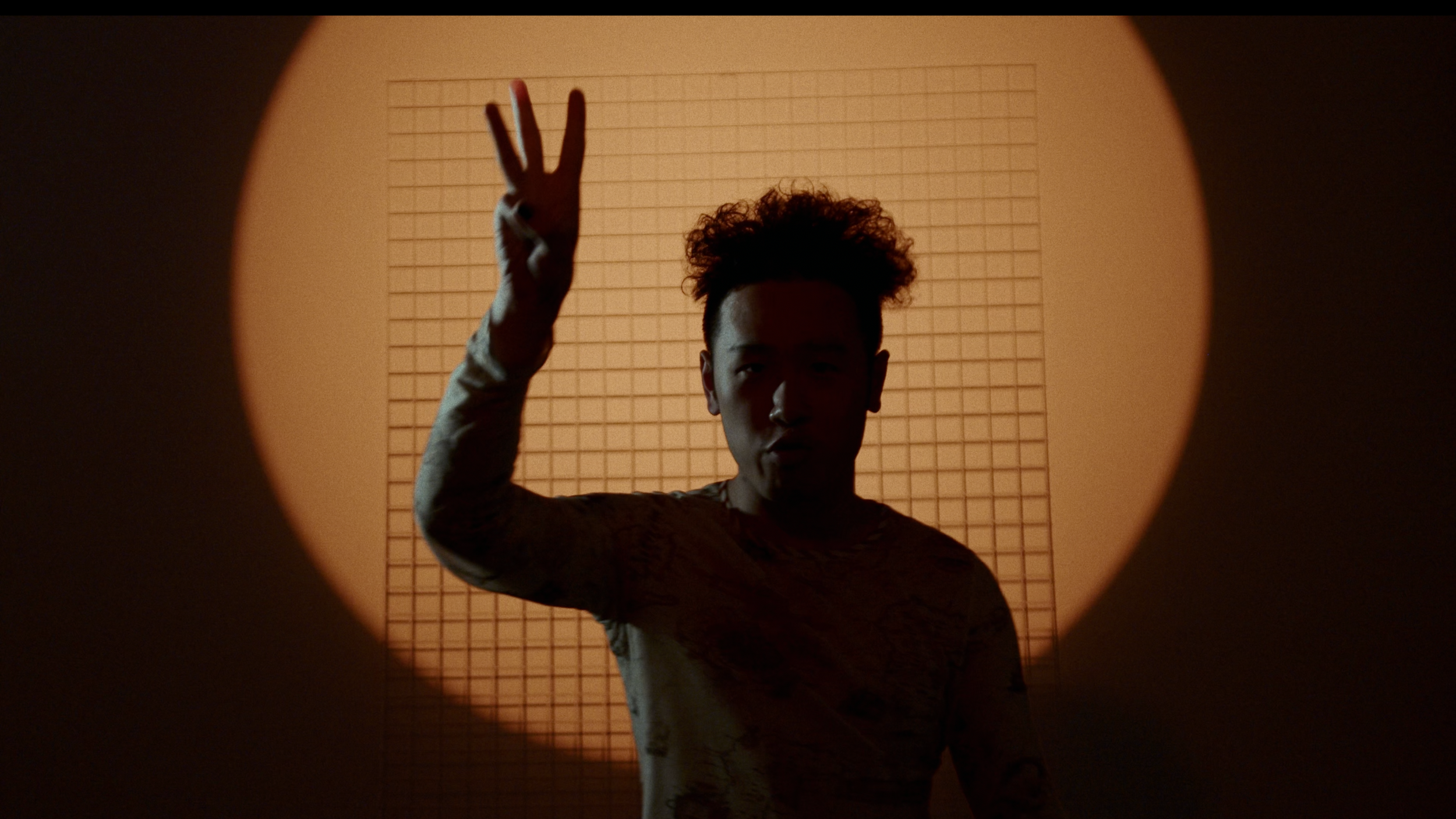A person with curly hair standing in front of a grid-patterned wall, raising their hand and making a peace sign.