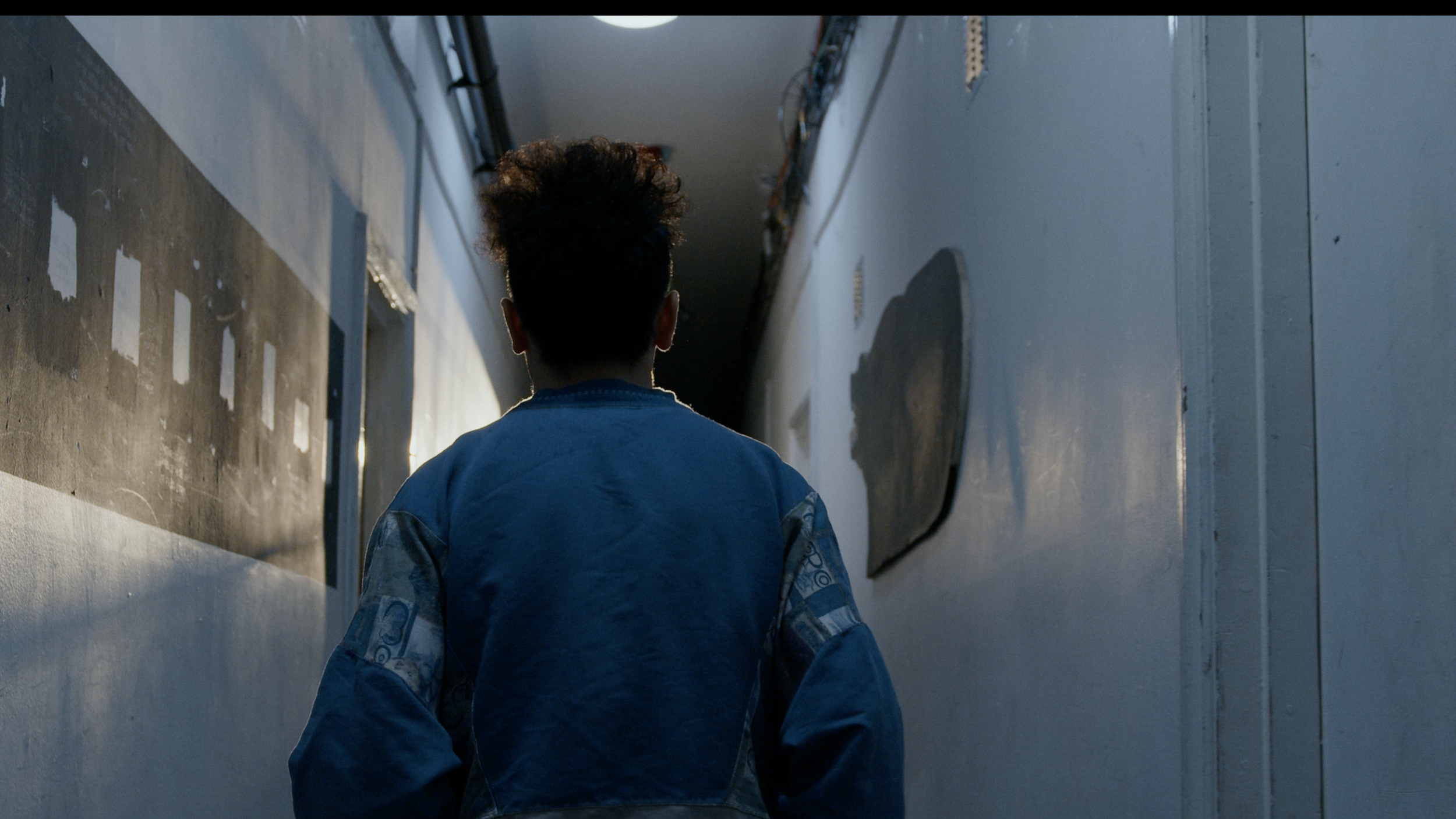 A person with curly hair walking down a dimly lit corridor with metallic and textured walls.