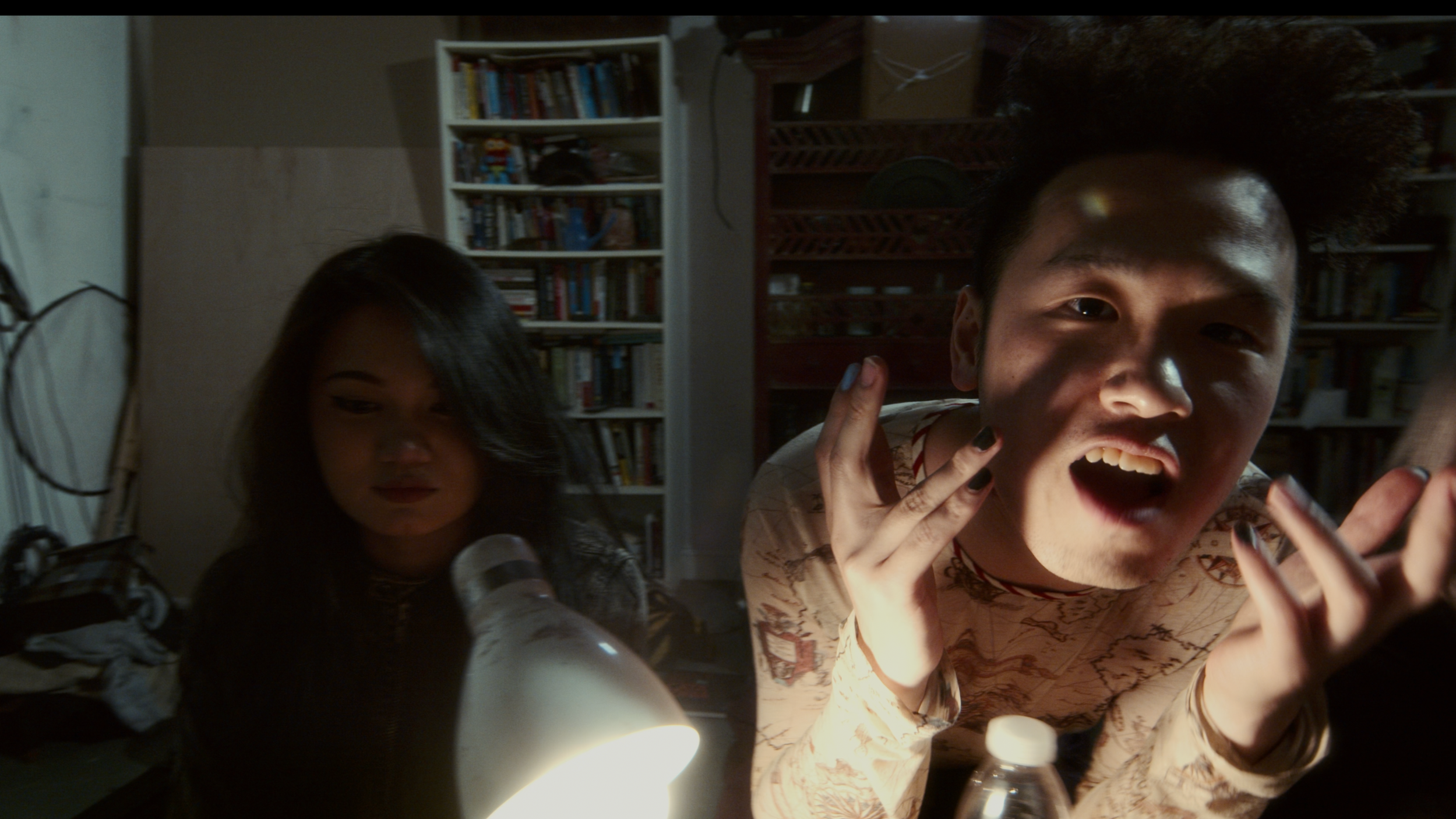 Two young people in a dimly lit room with bookshelves; one is a female looking down, and the other is a male leaning toward the camera with a surprised or expressive face, illuminated by a nearby desk lamp.