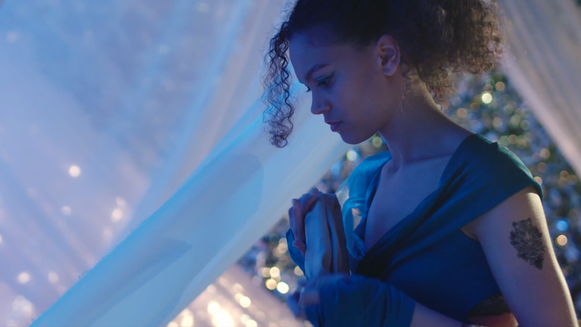 A woman with curly hair and a tattoo on her arm, standing with her hands clasped in prayer, surrounded by sheer fabric and bokeh lights.