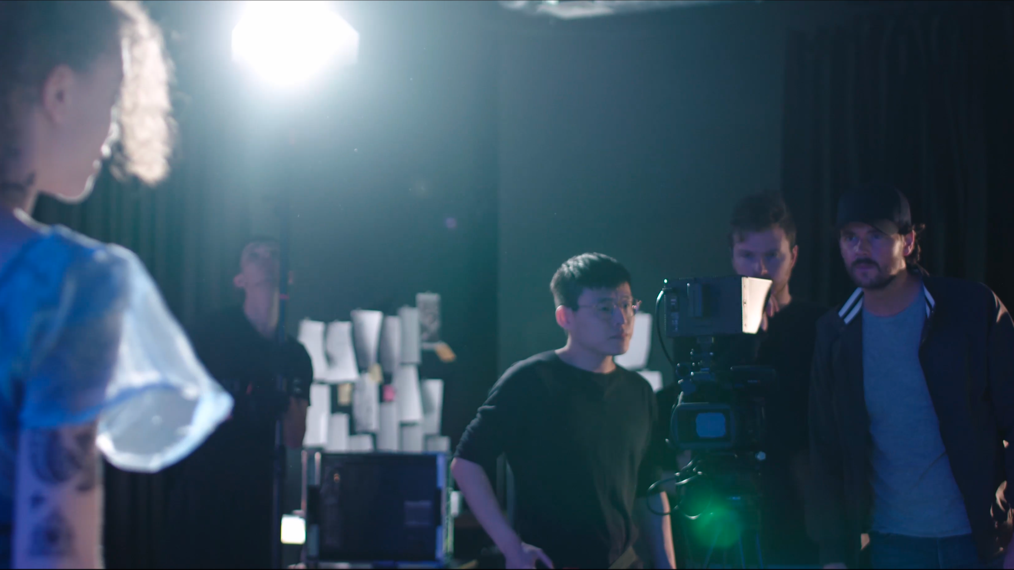 Film crew on set with camera and lighting, crew members focusing on shot, background with woman standing and paper cups on table, dark backdrop