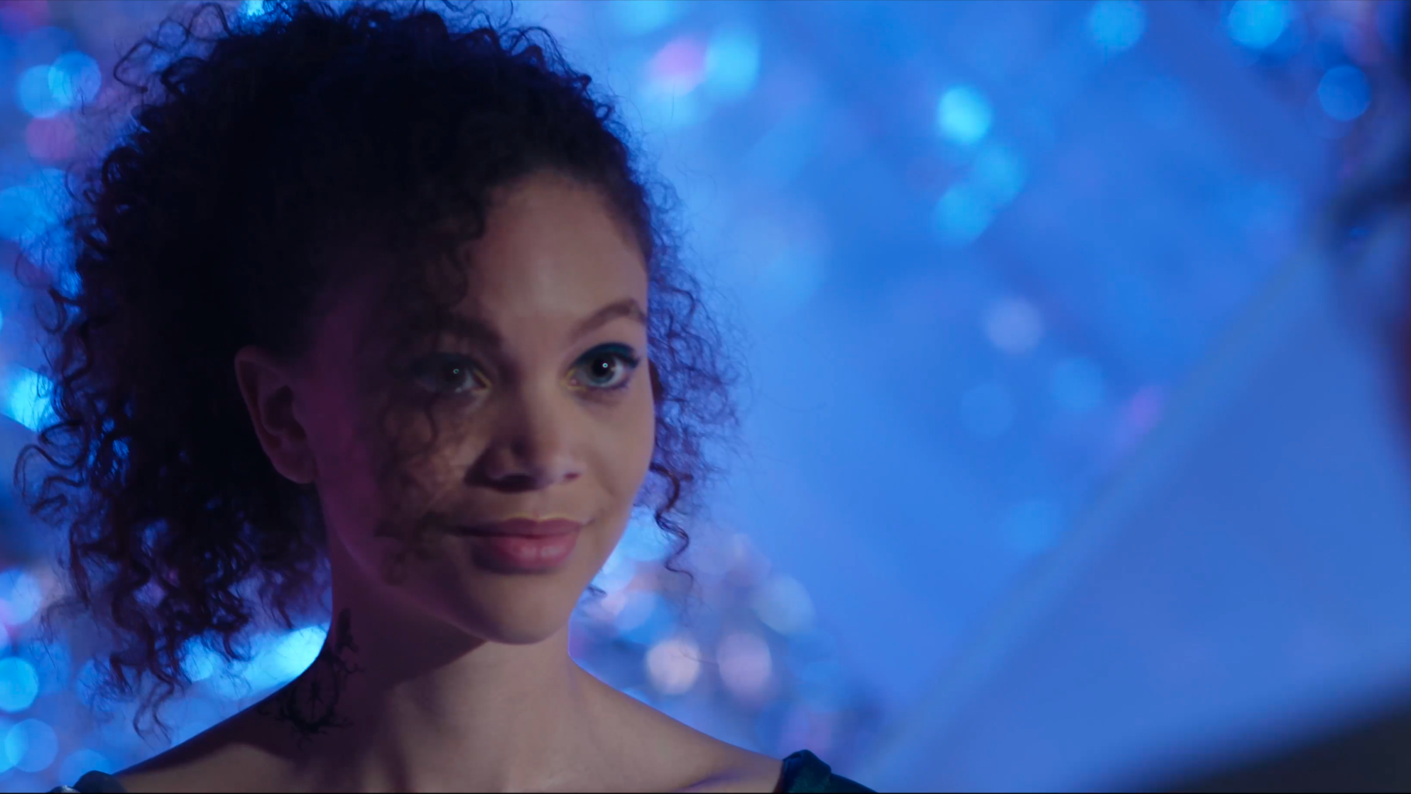 A woman with curly black hair and light skin, speaking to someone out of the frame, with a shimmering blue background and blurred colorful lights.