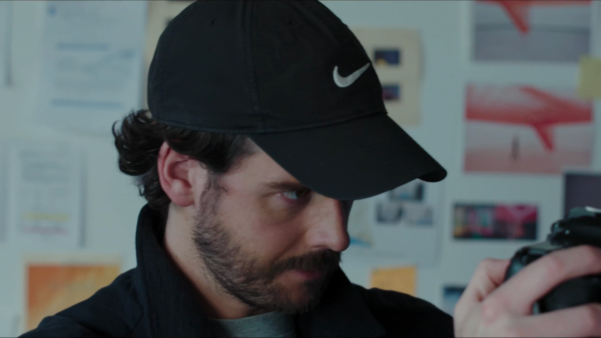 Close-up of a man wearing a black Nike cap looking at a camera, with a background of bulletin boards and papers on a wall.