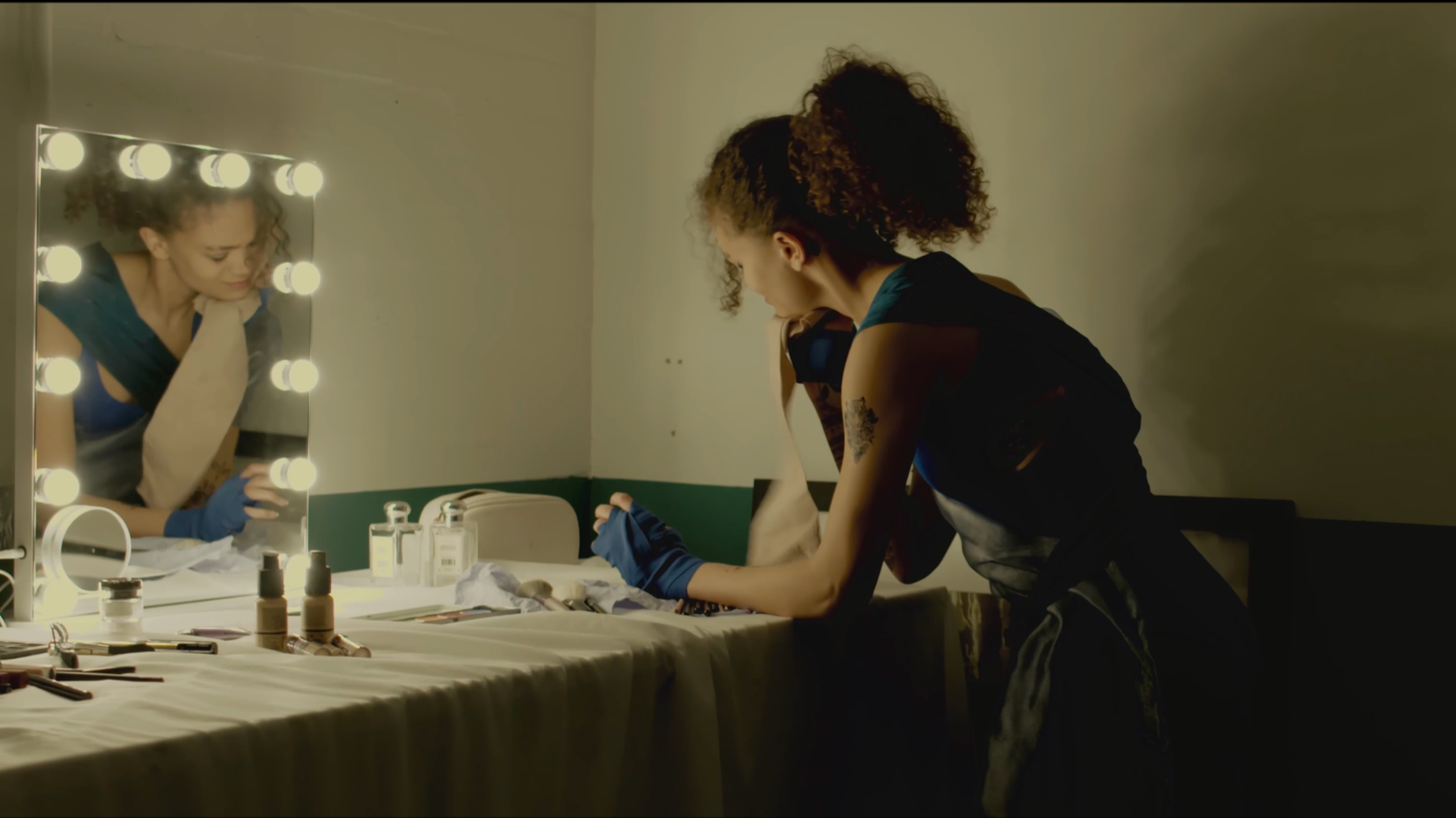 A woman getting ready in front of a brightly lit mirror, with beauty products and makeup tools on a table, in a dimly lit room.