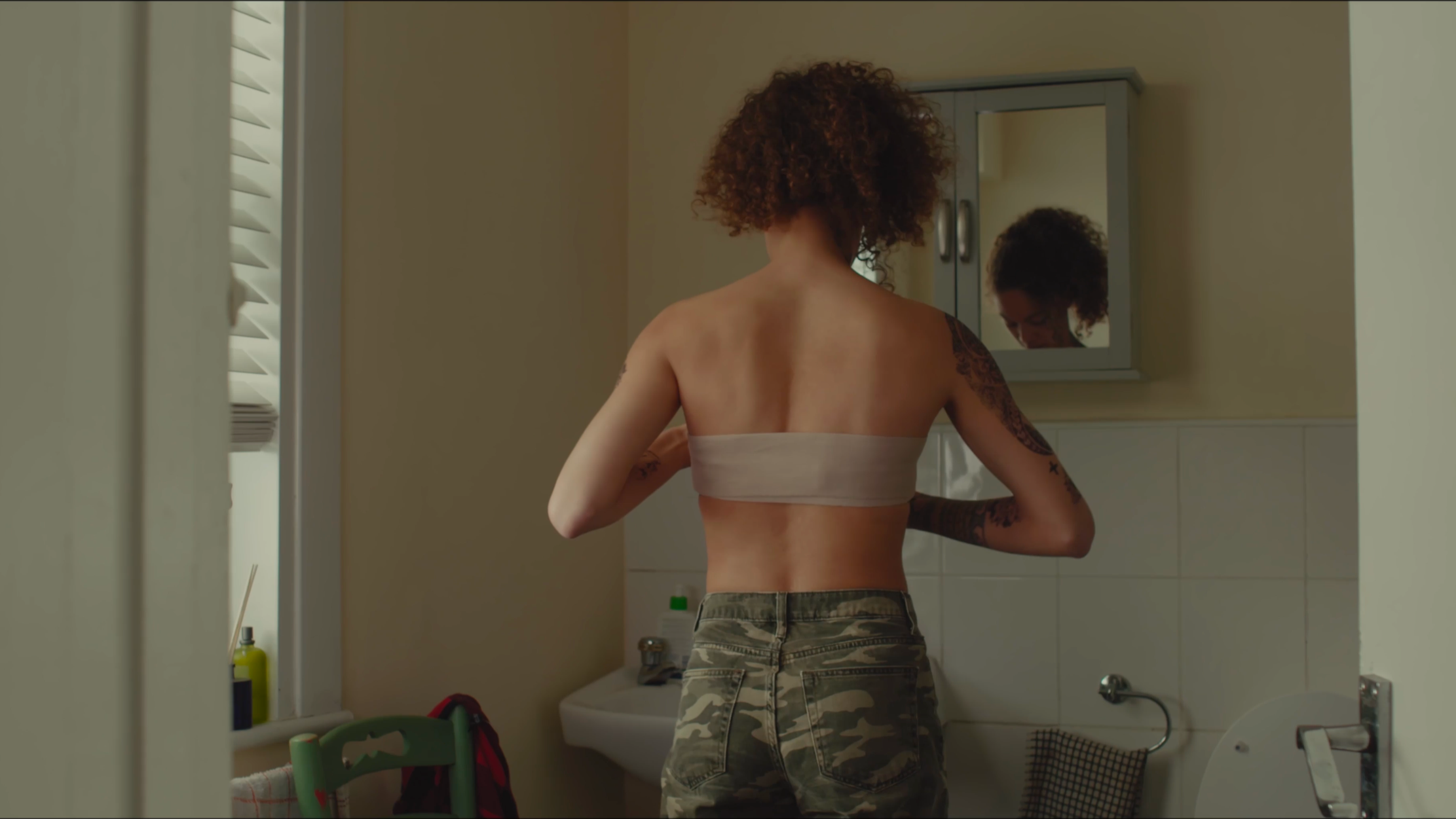 A woman with curly hair and tattoos on her arms stands in front of a bathroom mirror, adjusting her beige strapless top, wearing camouflage pants, with a bathroom sink and window visible in the background.