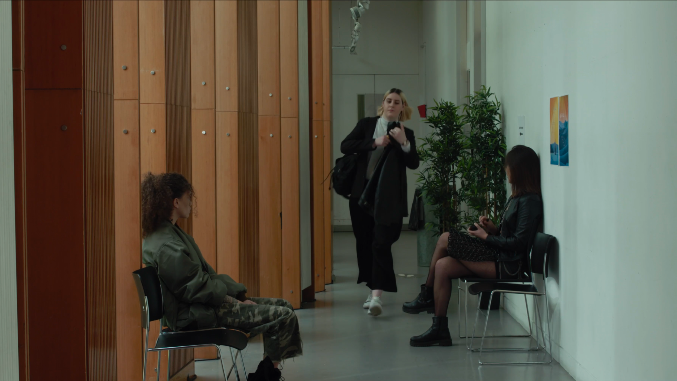 Three women are sitting on black chairs in a waiting area with a white wall and green plants; a woman in a blazer is walking past holding a jacket.