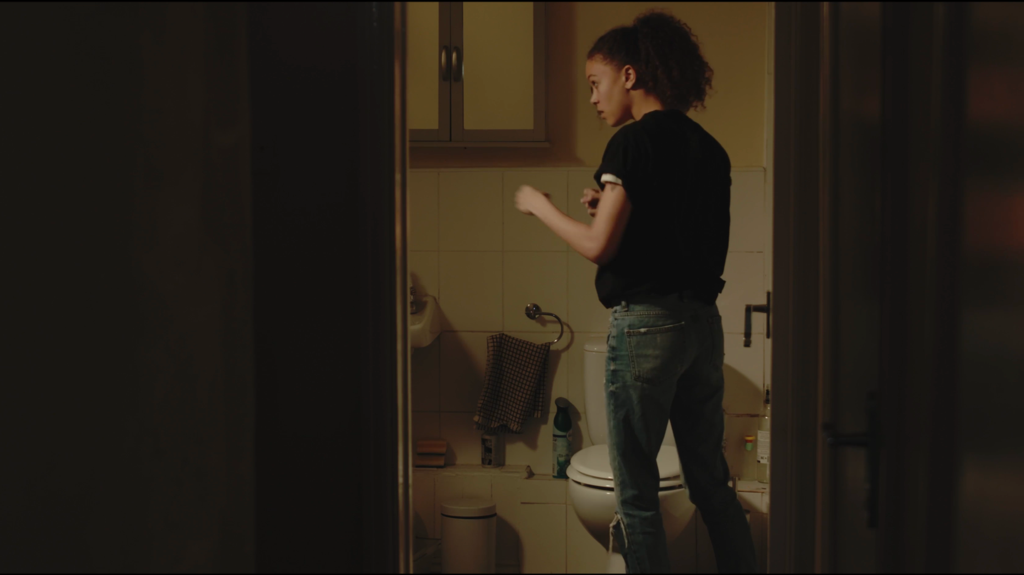 A young woman with curly hair standing in a bathroom and looking at herself in a mirror.
