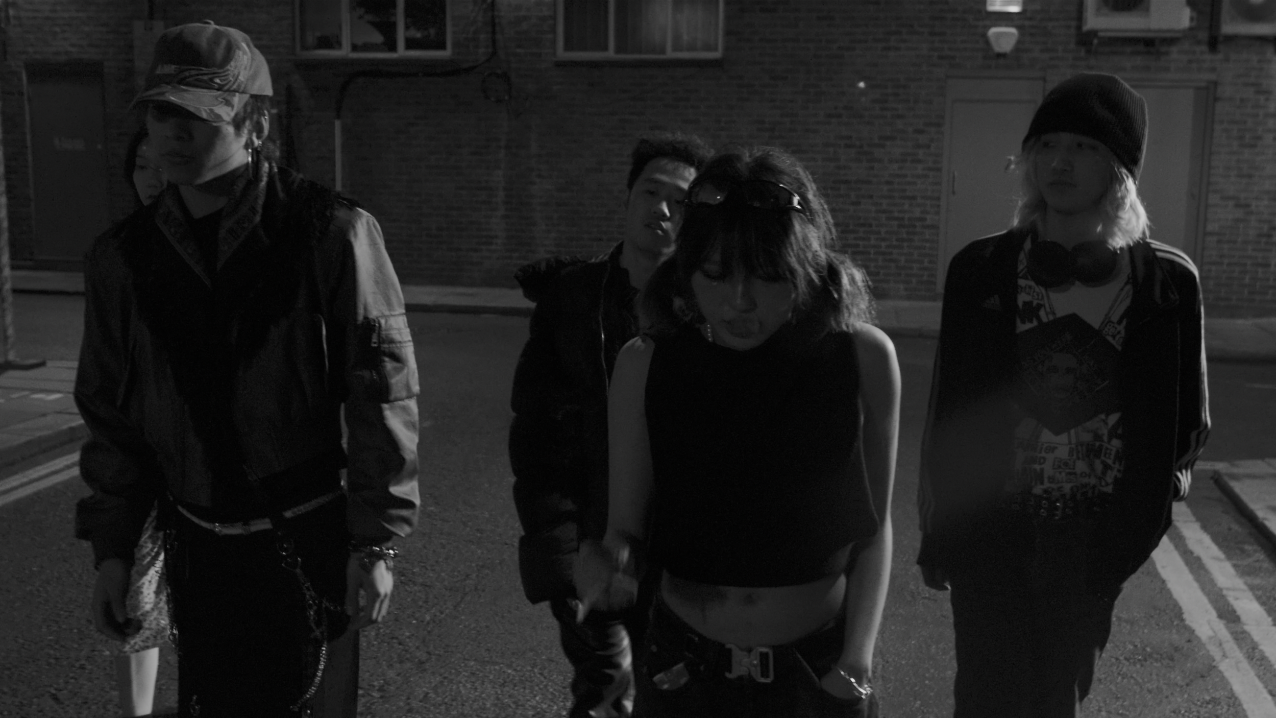 Group of five young people walking together at night on a city street, dressed in casual, edgy clothing with accessories like hats, sunglasses, and headphones.
