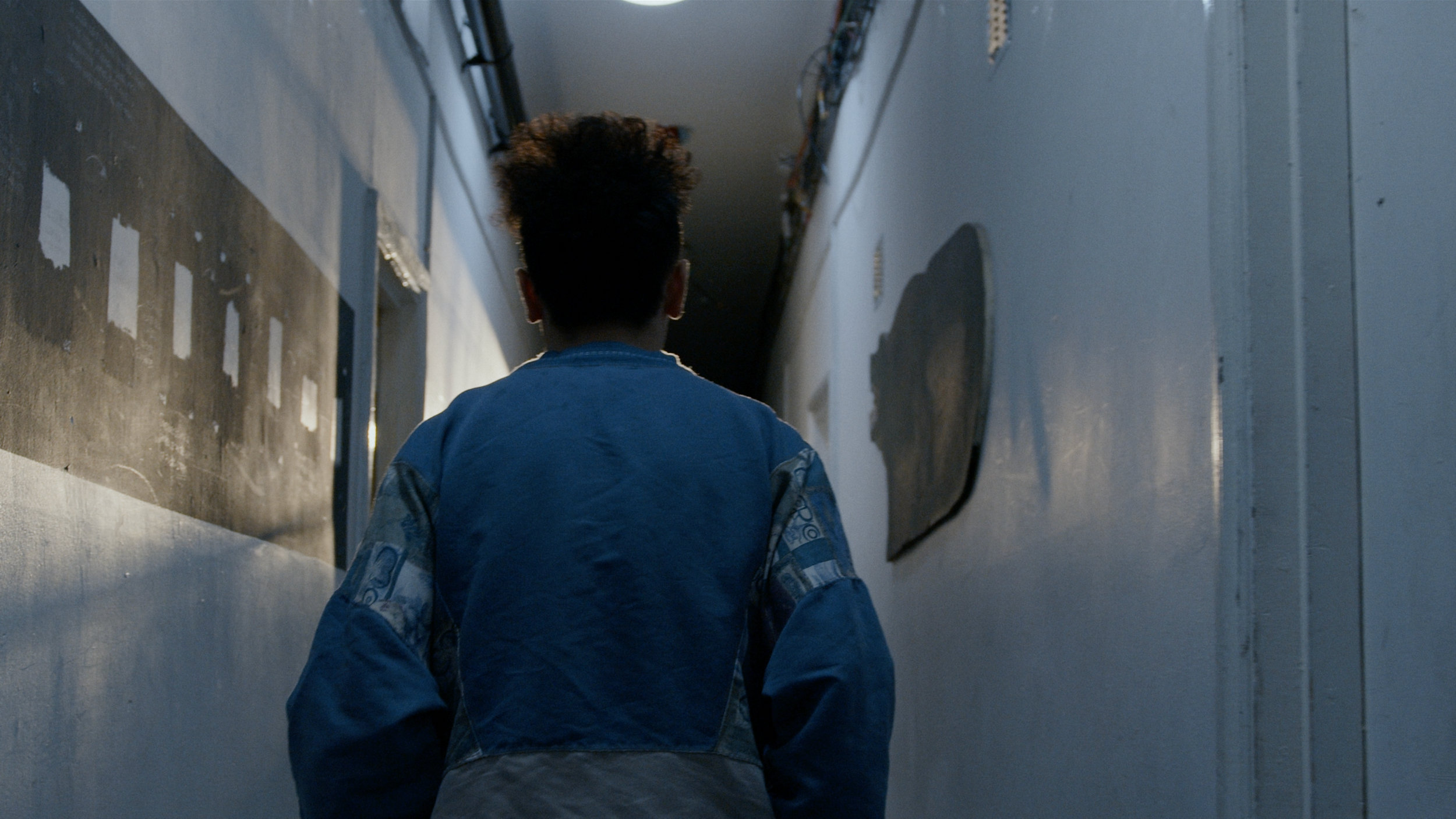A person with short dark hair wearing a blue jacket walking down a narrow, dimly lit corridor with unfinished walls and exposed wiring.