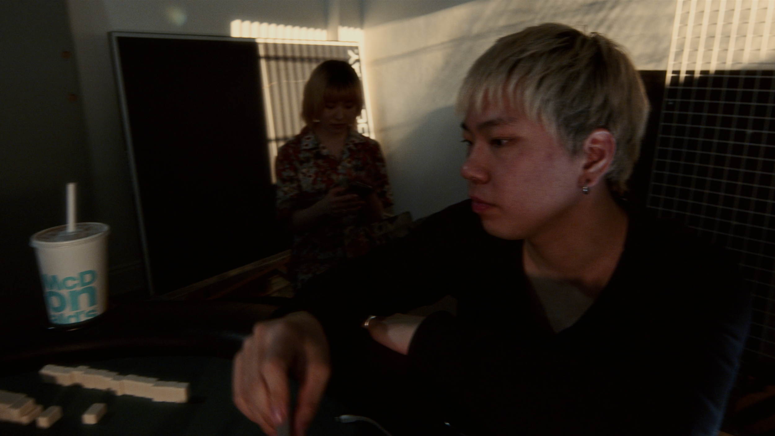 A person with short blond hair and earrings is sitting at a table in a dimly lit room, looking at a device. In the background, another person with shoulder-length hair wearing a floral shirt is using a mobile phone. There is a McDonald's cup on the t