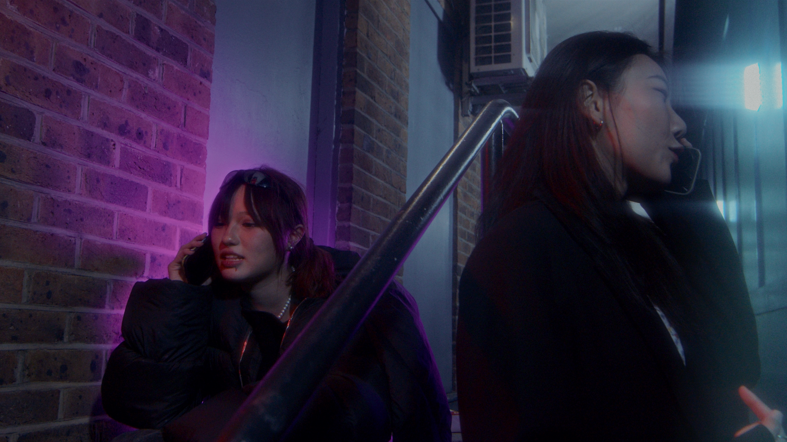 Two women are sitting against a brick wall, talking on their phones. One woman has dark hair and is wearing black, and the other has purple hair and is also dressed in dark clothing. The scene appears to be at night with colorful lighting.