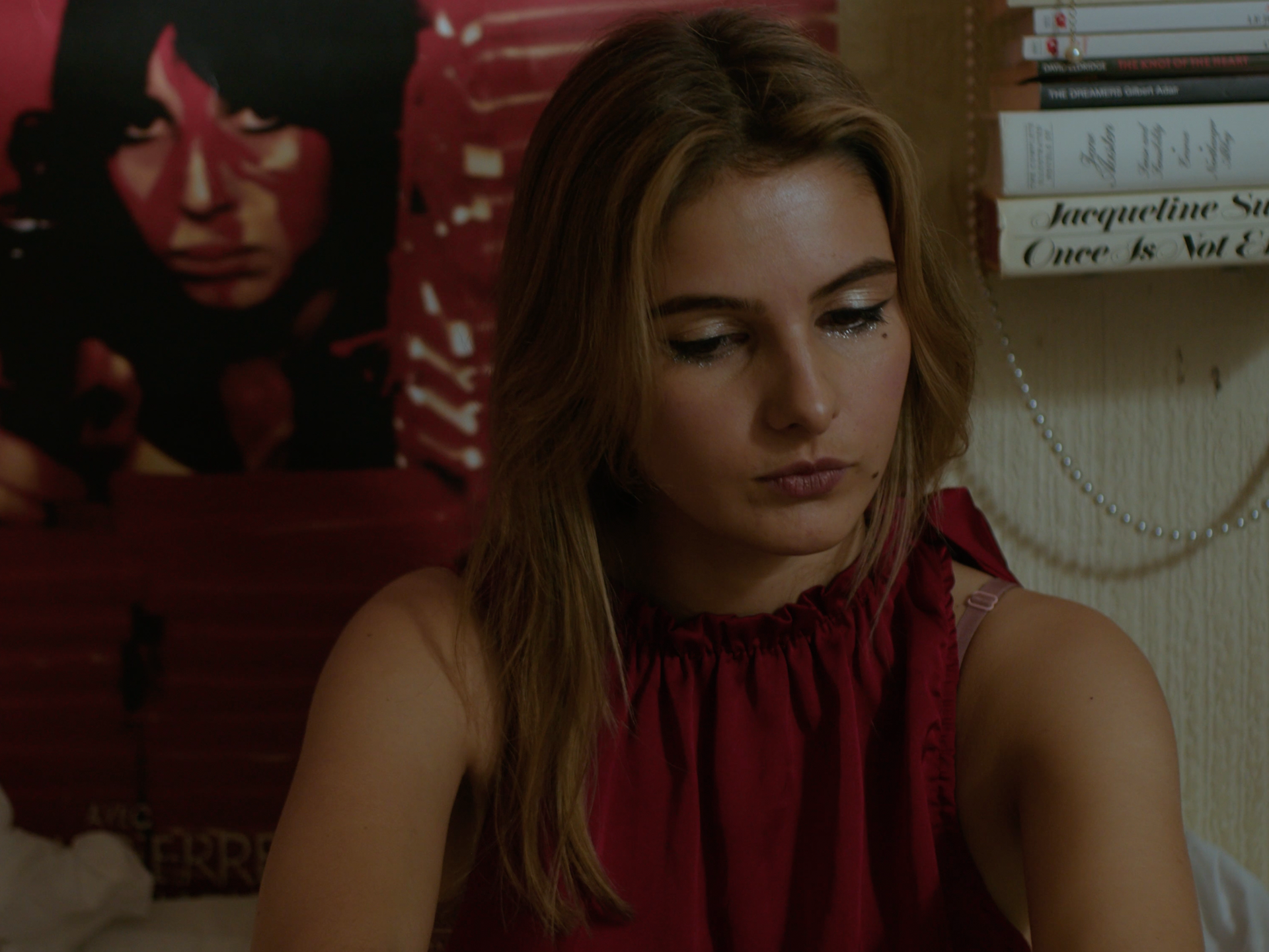 A young woman with long, light brown hair looking down with a serious expression, wearing a red sleeveless top, in a room with a white wall, books on a shelf, and a large poster of a woman's face with dark hair in the background.