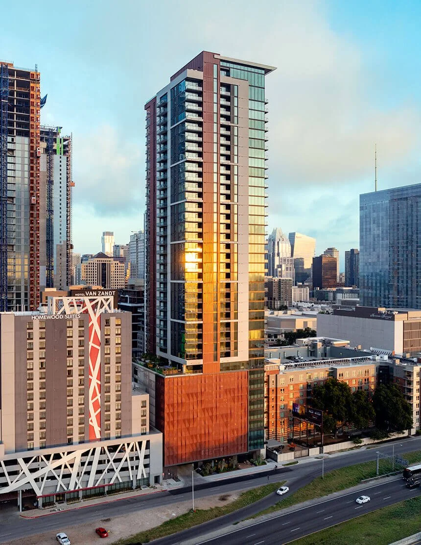 A tall modern high-rise building with glass and metal exterior, reflecting the sunset, located in an urban area with other high-rise buildings around, and a road with cars in the foreground.
