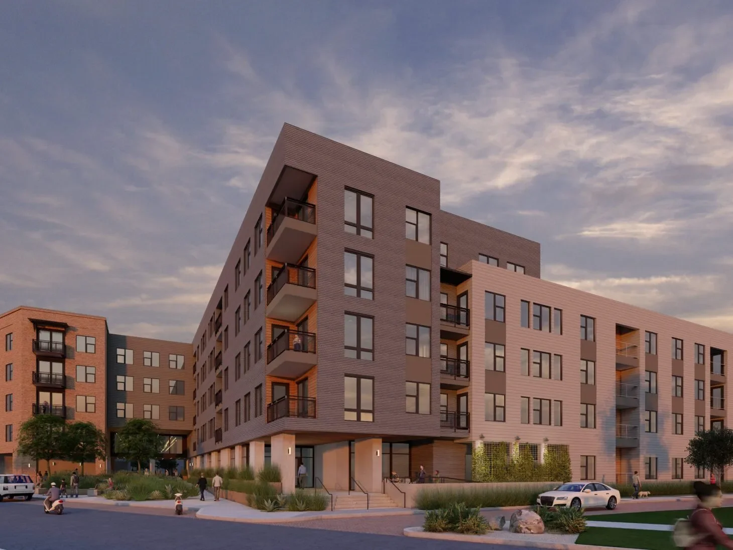 A modern multi-story apartment building with brick and beige siding, several balconies, and large windows, set against a sunset sky. There are people walking, trees, and parked cars near the building.