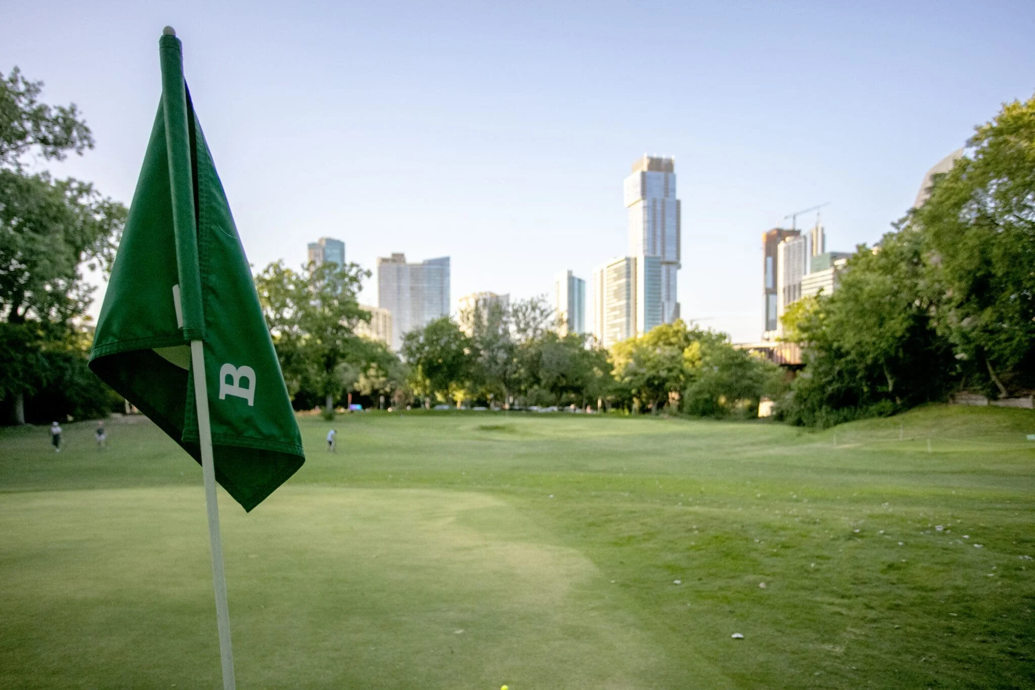 A golf course with a green flag in the foreground and a city skyline in the background.