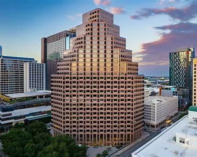 A cityscape featuring a large stepped office building with a pinkish-brown facade, surrounded by other modern buildings and a cloudy sky during sunset.