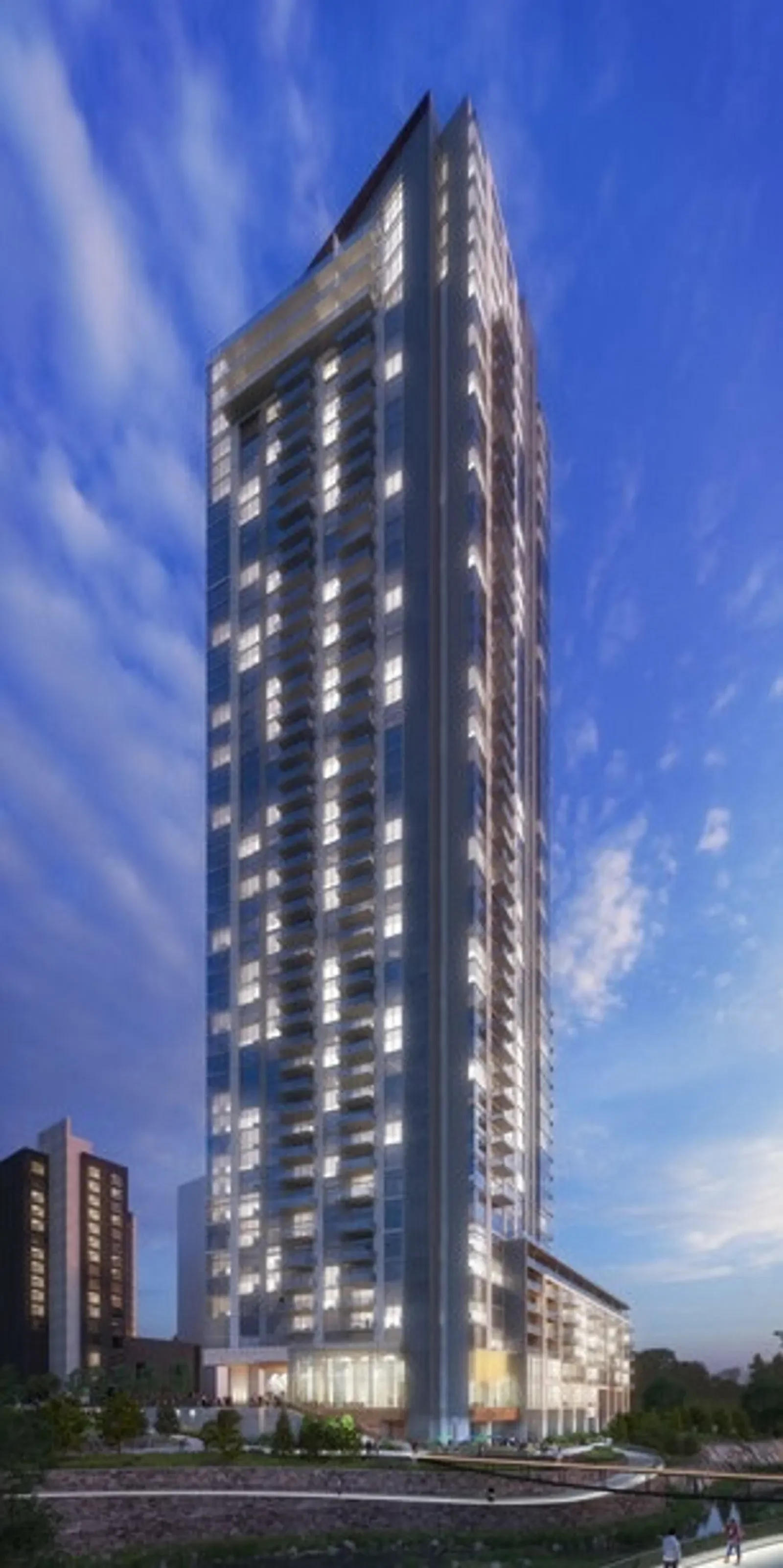 A tall modern skyscraper with glass balconies and windows, illuminated at the base, set against a blue sky with scattered clouds.