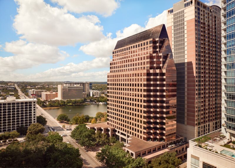 A tall modern office building with a glass facade reflecting the sky, situated in an urban cityscape near a river, with other high-rise buildings and trees in the surrounding area.