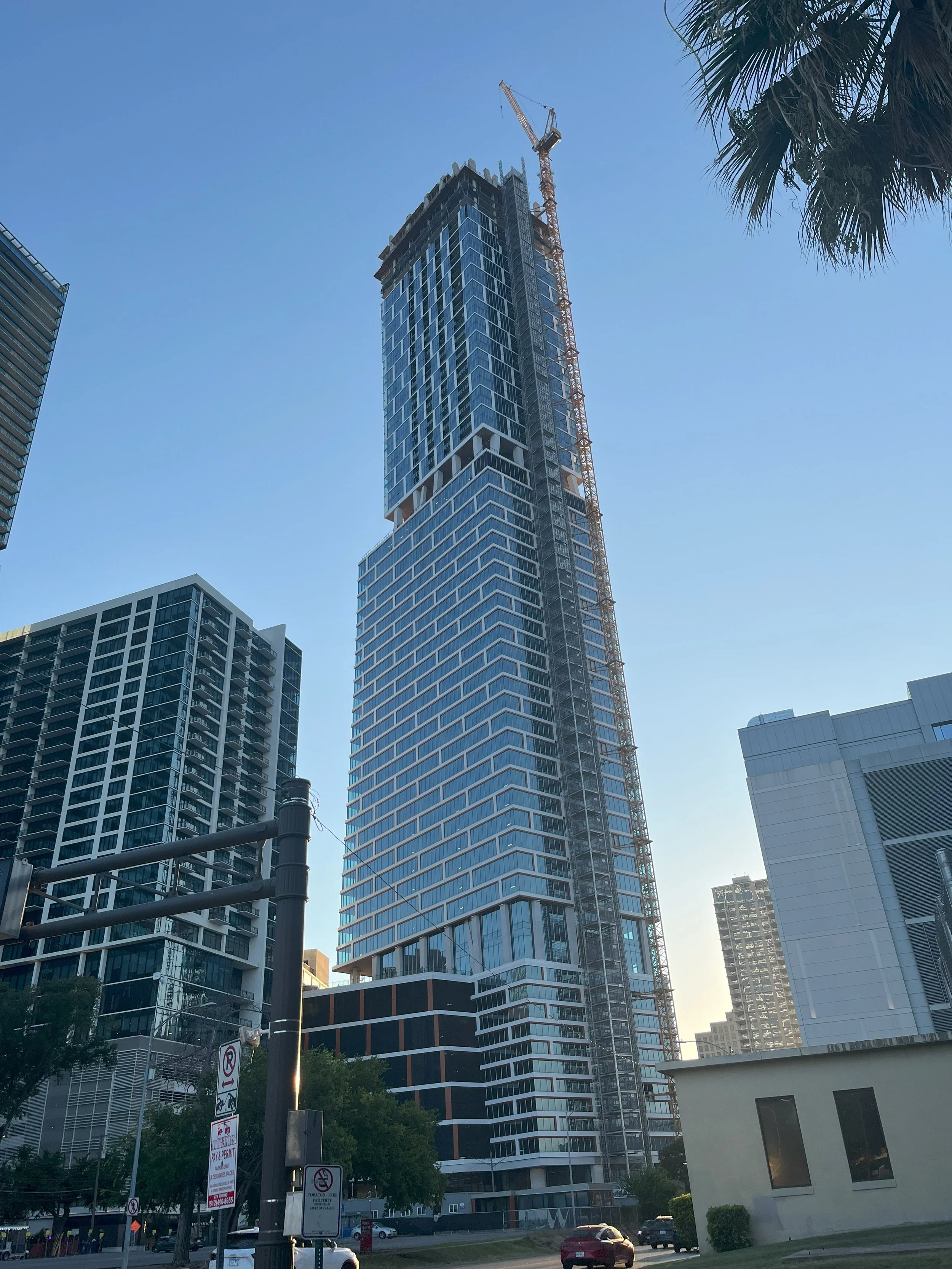 A tall modern glass skyscraper under construction in an urban area, with a crane on top and other high-rise buildings around it, against a clear blue sky.