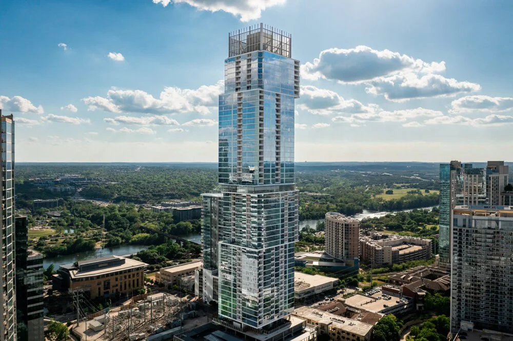 Tall modern glass skyscraper in a cityscape with green parks and a river in the background under a blue sky with clouds.
