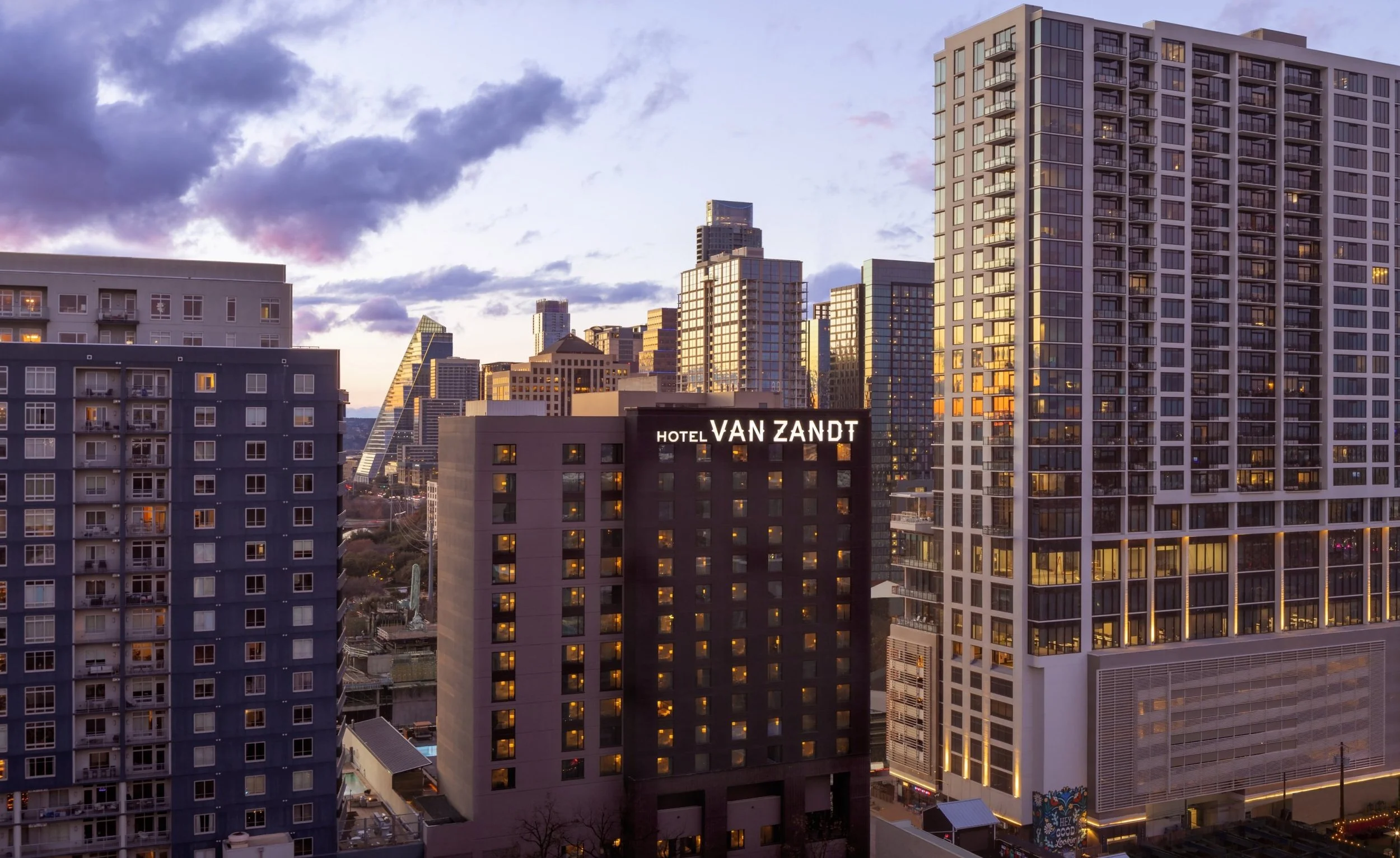 City skyline at sunset with tall buildings, including a hotel named 'Hotel Van Zandt' in the foreground.