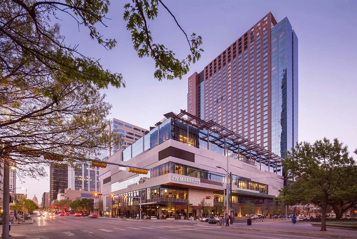 Nighttime cityscape featuring a modern building with JW Marriott signage at street level, surrounded by trees, cars, and pedestrians with a purple sky background.