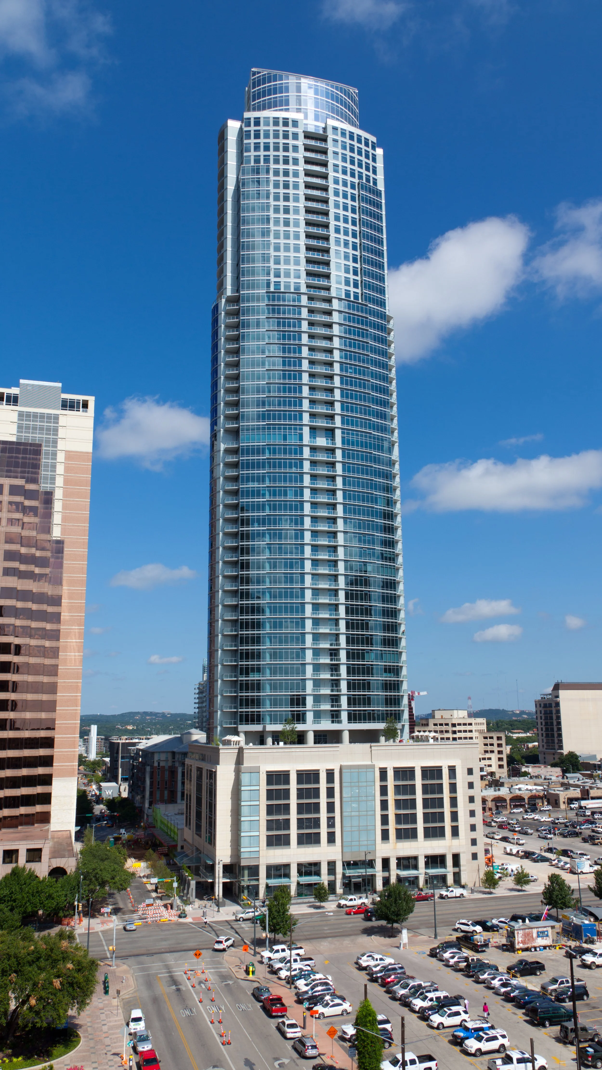 Tall modern skyscraper in an urban downtown area with parking lot and busy street in foreground and partly cloudy blue sky.