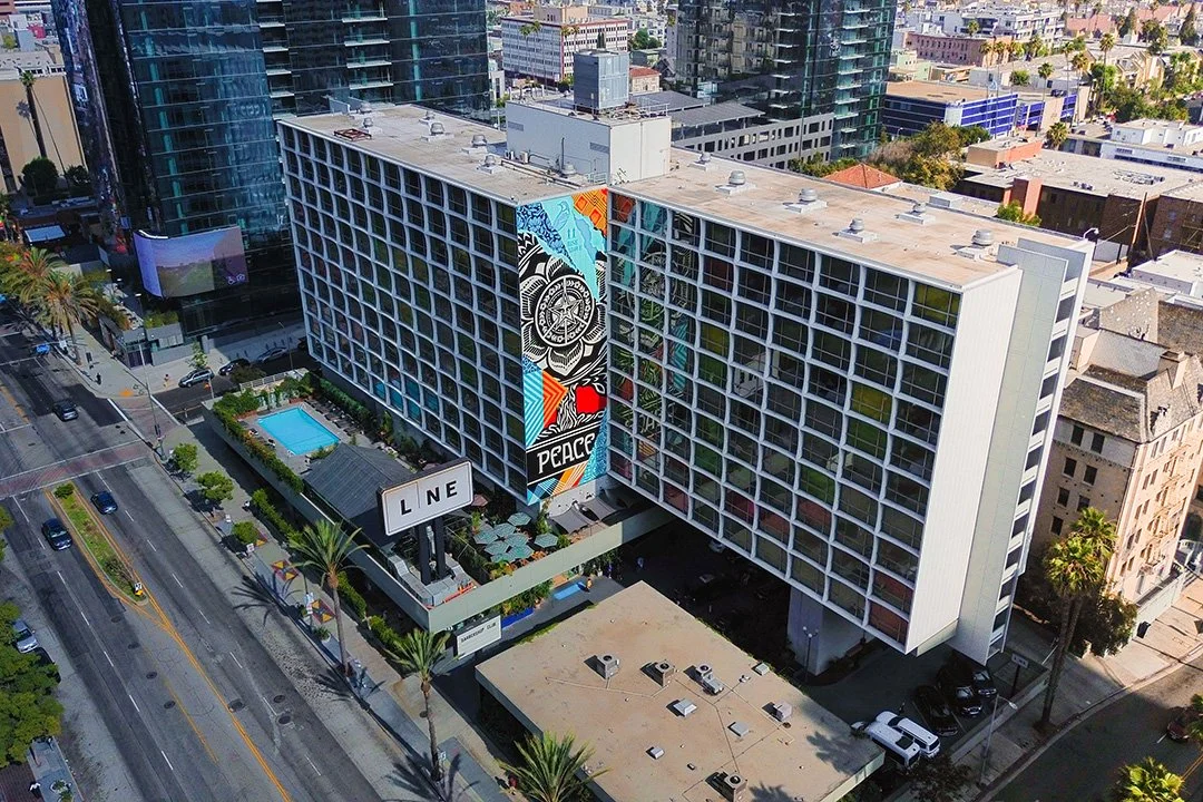 Aerial view of city of Austin block featuring a large multi-story hotel with a colorful mural on its side, a swimming pool with lounge chairs, and palm trees lining the street.